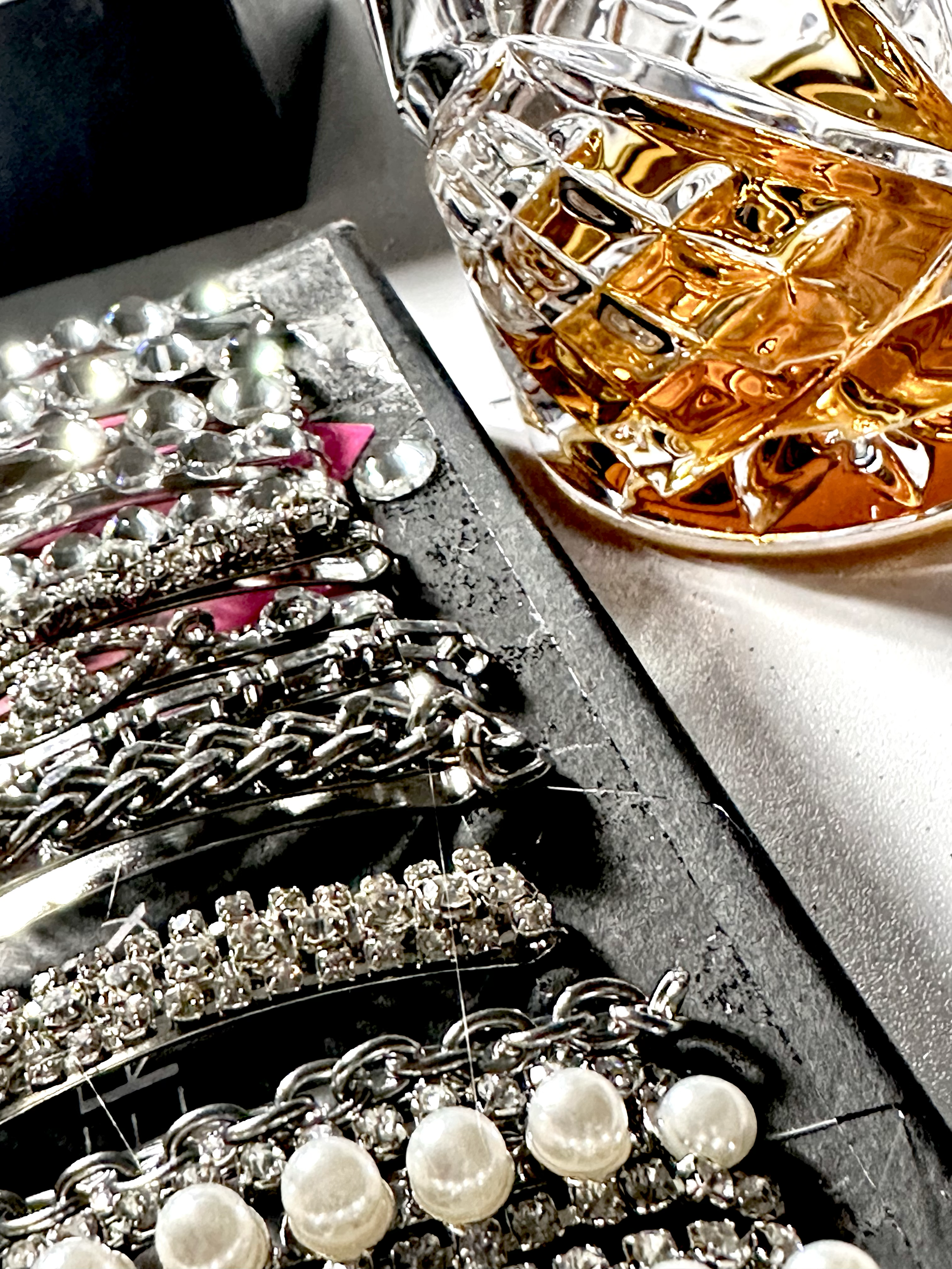 Close-up of jewelry and a glass of whiskey. The jewelry includes pearl necklaces, rhinestone bracelets, and other silver-toned pieces, placed on a black jewelry stand. A glass of whiskey with ice is visible in the background.
