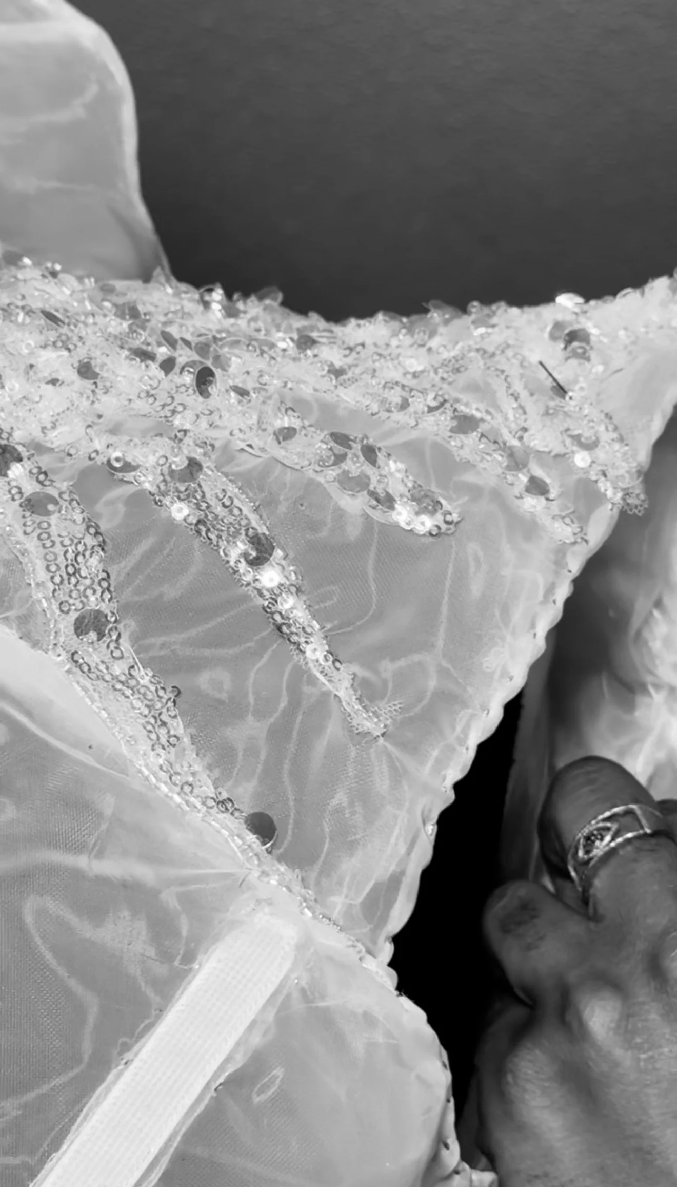 Close-up of a hand wearing a ring, grasping a fabric adorned with sequins and beads, possibly part of a wedding dress or formal gown.
