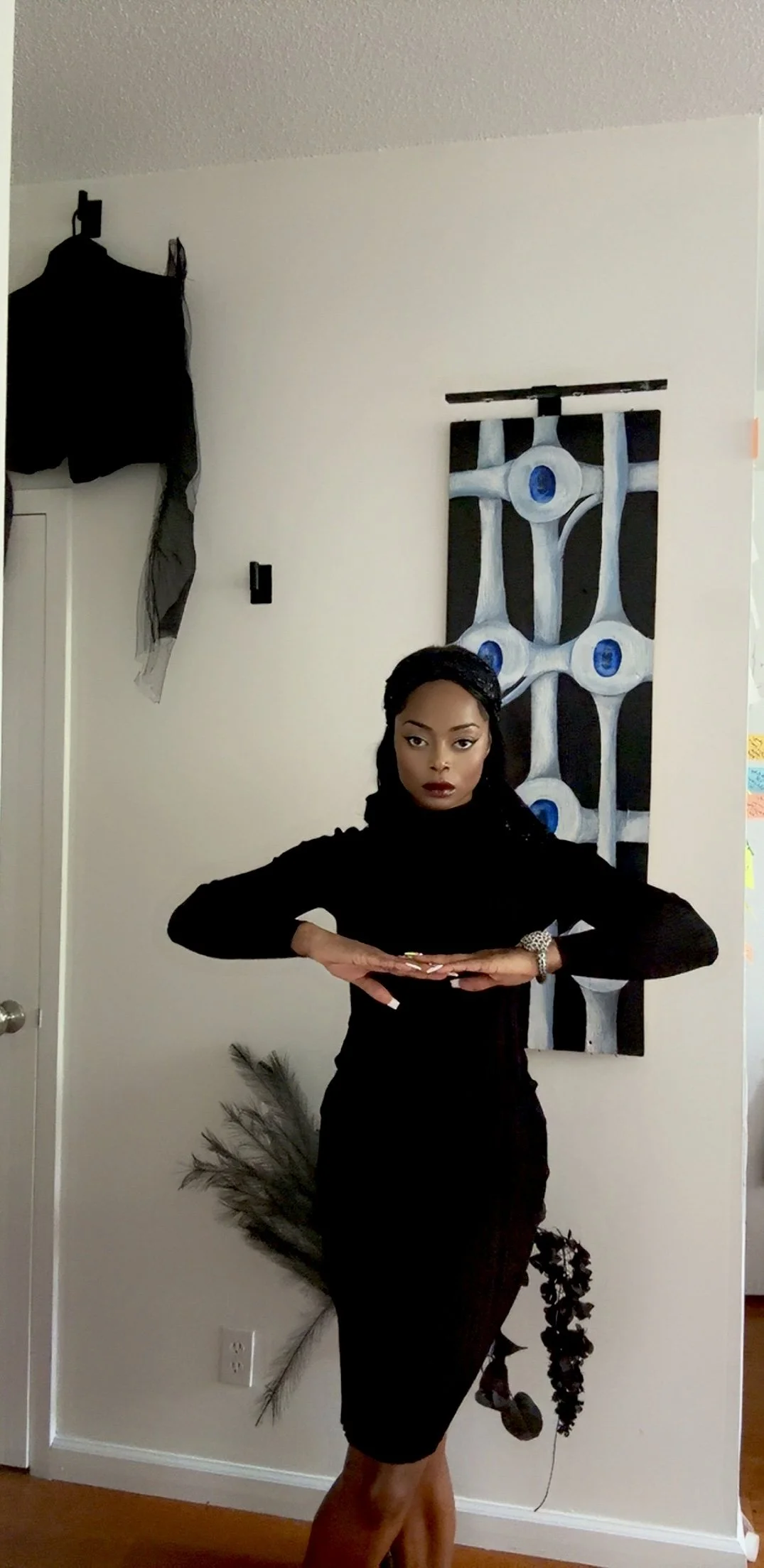A woman dressed in black with dark lipstick and makeup, posing indoors in front of a white wall with an abstract blue, white, and black painting. She has her hands together in front of her chest, with a peacock feather visible at her side.