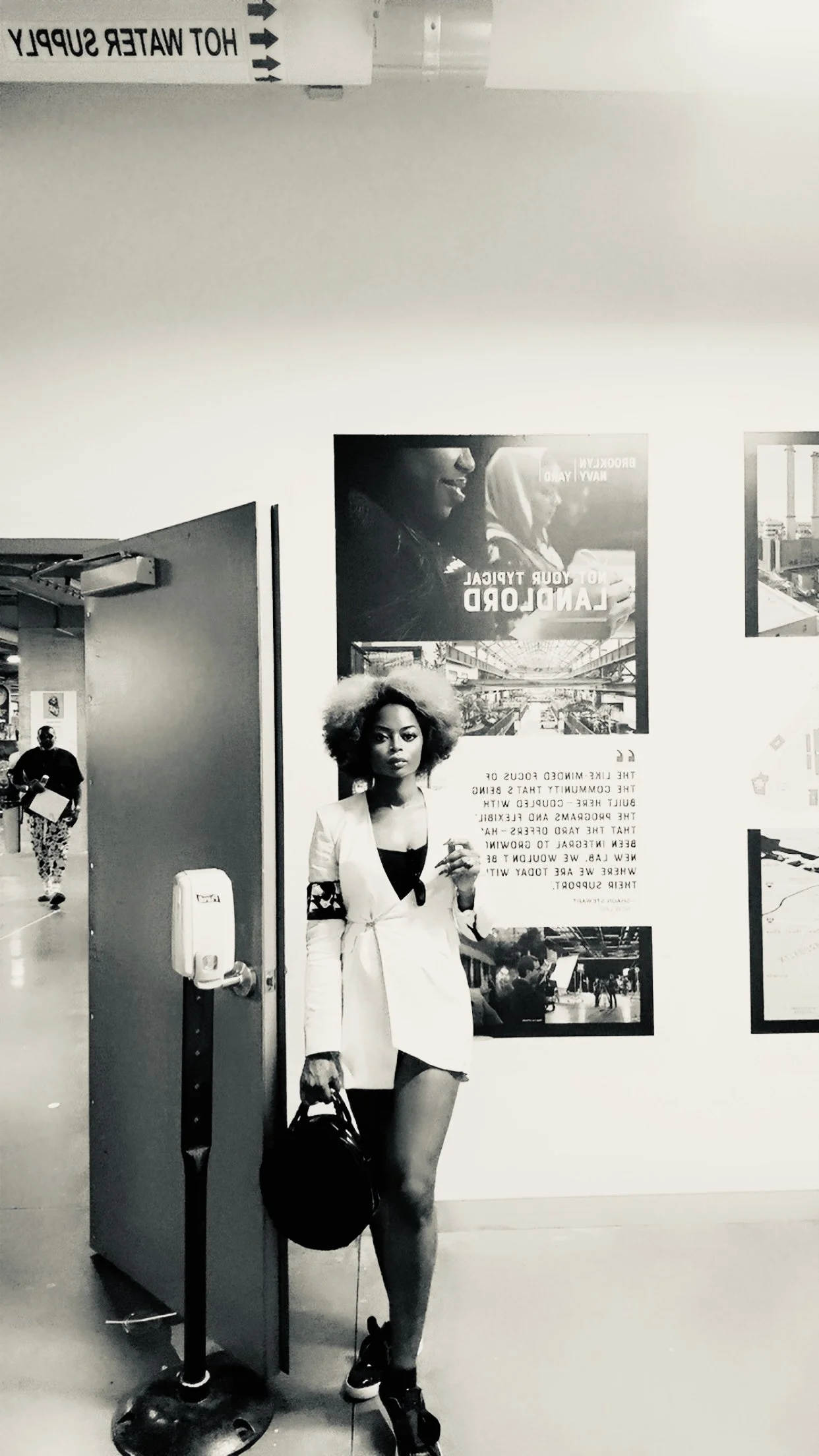 A woman with curly hair standing next to a wall with posters, holding a black bag while wearing a white blazer and shorts, inside an airport.