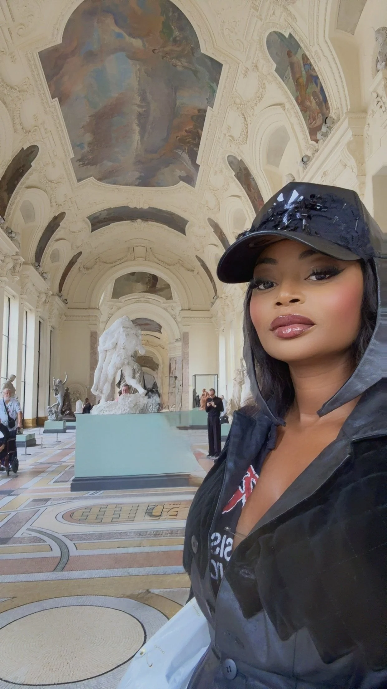 A woman with makeup and a black shiny hat takes a selfie inside a grand, decorated museum or gallery with sculptures and detailed ceiling paintings.