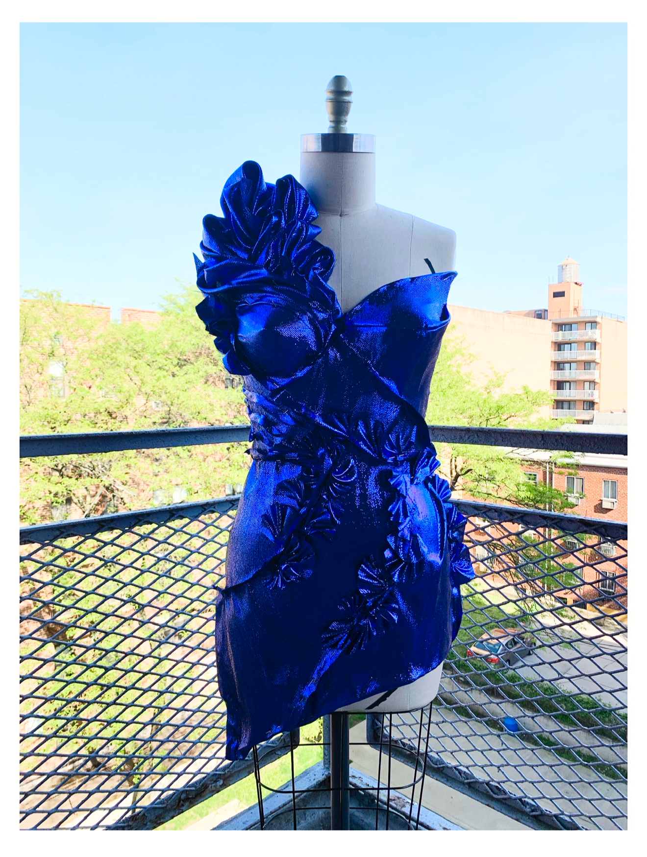 A dress on a mannequin features a vibrant blue, ruffled, and textured fabric with floral details, displayed on a balcony with a cityscape background.