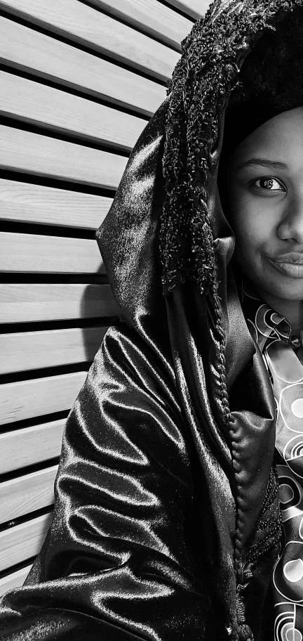 A young girl with braids and a shiny jacket, partially visible, standing in front of a wooden slat wall.