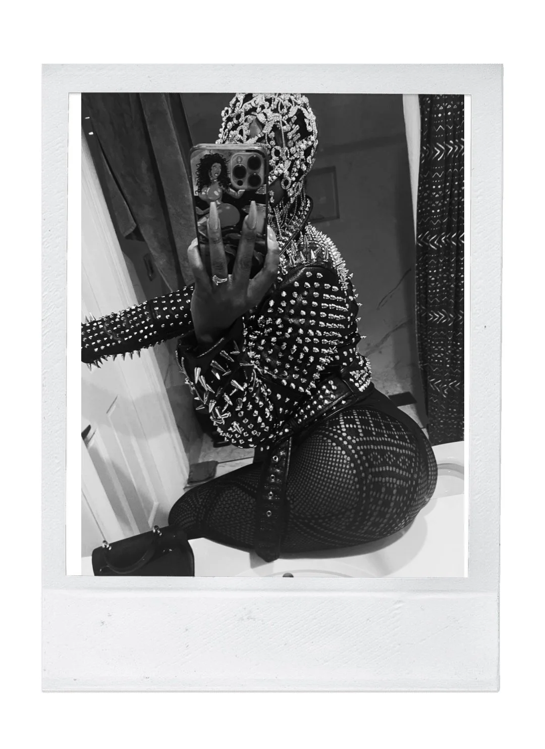 A person taking a mirror selfie with a phone, wearing a spiked studded jacket and fishnet leggings, in a room with curtains.