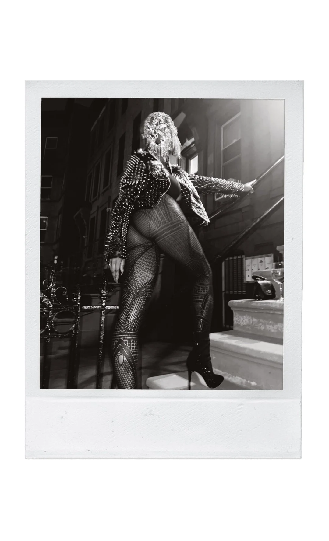 Black and white photograph of a woman wearing high heels, patterned tights, and a studded leather jacket, standing on stairs in front of buildings, with sunlight coming from the right.
