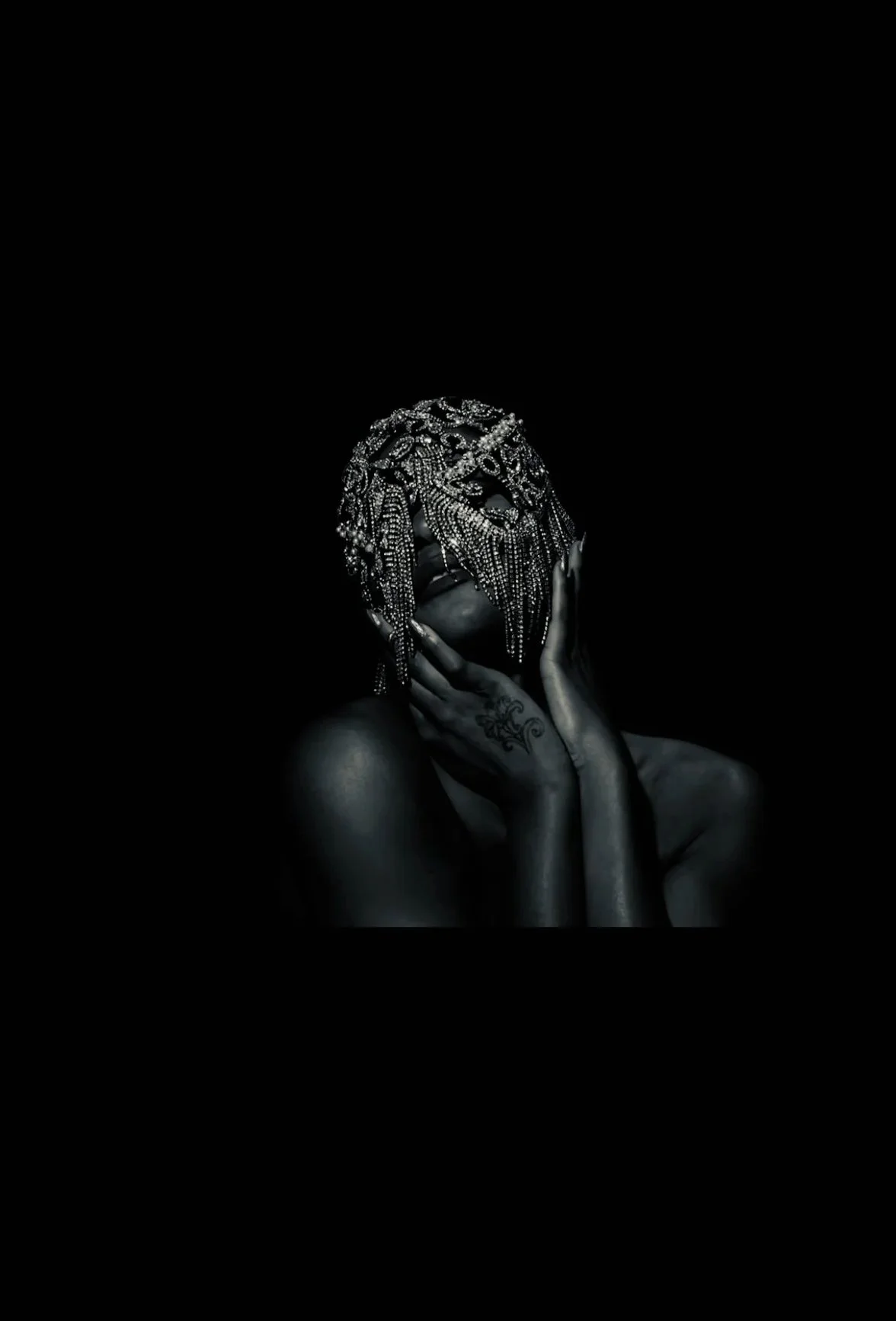 A dark image of a person with dark skin, partially nude, holding their face with their hands. They are wearing an intricate beaded headpiece or mask that covers their face, featuring a mask-like design with hanging chains or beads.