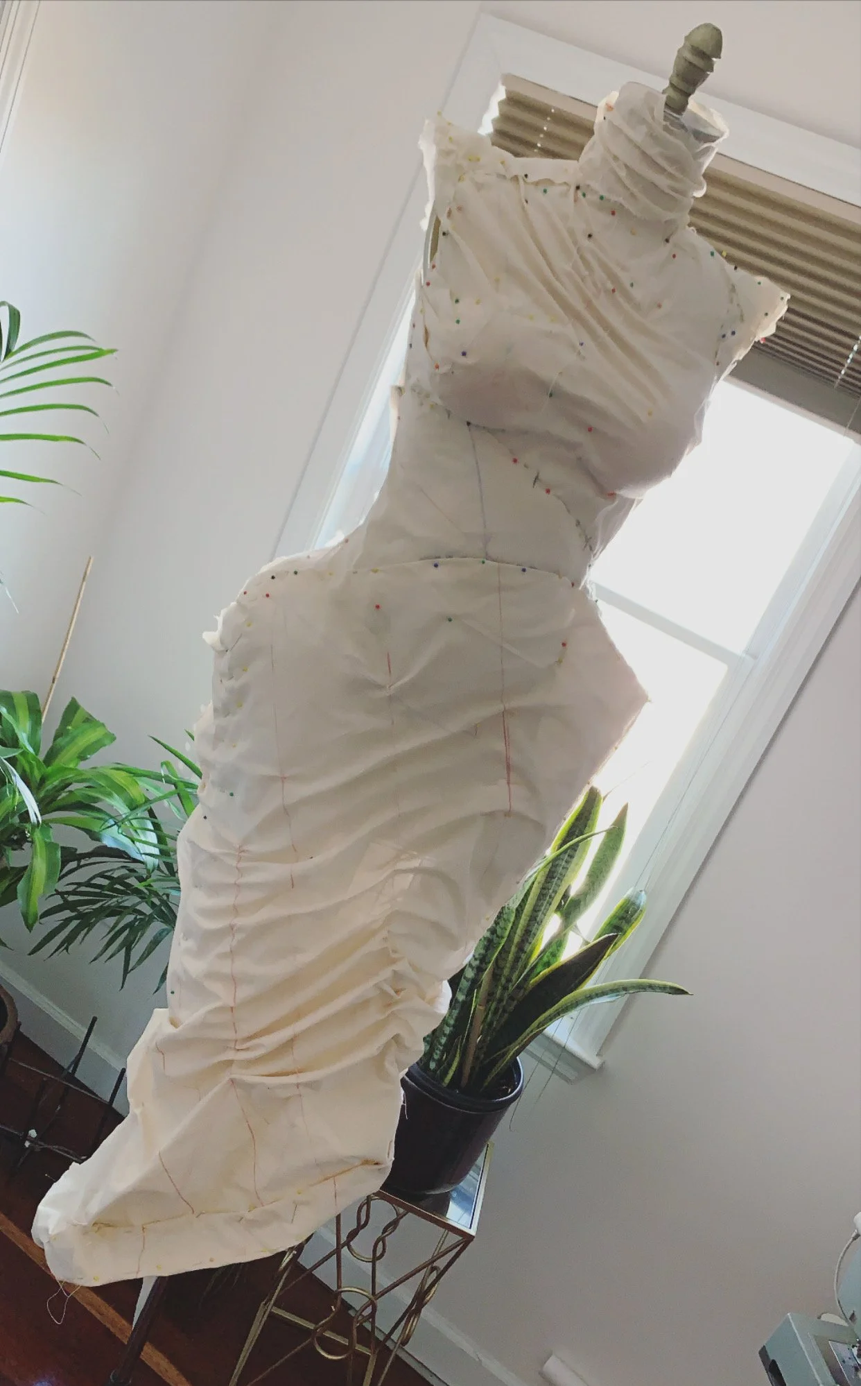 Partial view of a dress form mannequin covered in white fabric with sewing pins, positioned near a window with blinds, with potted plants nearby.