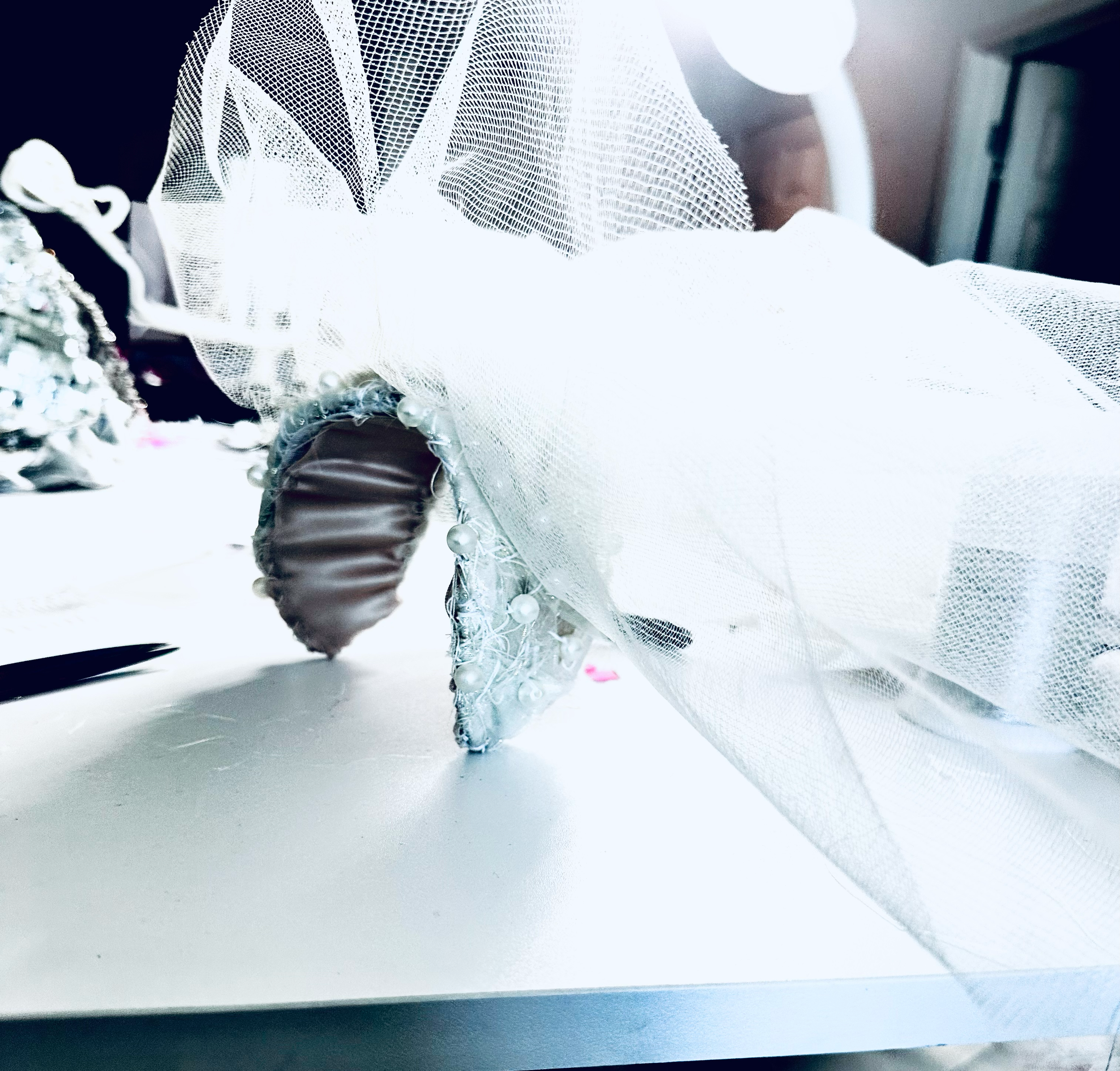 Close-up of a decorated high heel shoe with pearls and beads, partially covered by a white veil or tulle fabric.