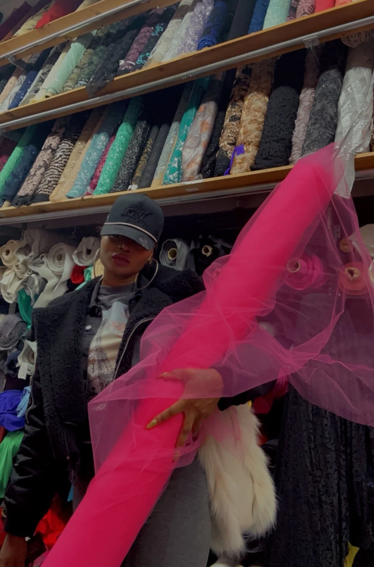 Woman standing in front of shelves of fabric rolls in a store, wearing a black cap, black jacket, and a patterned shirt, with pink tulle fabric and white fur in the foreground.