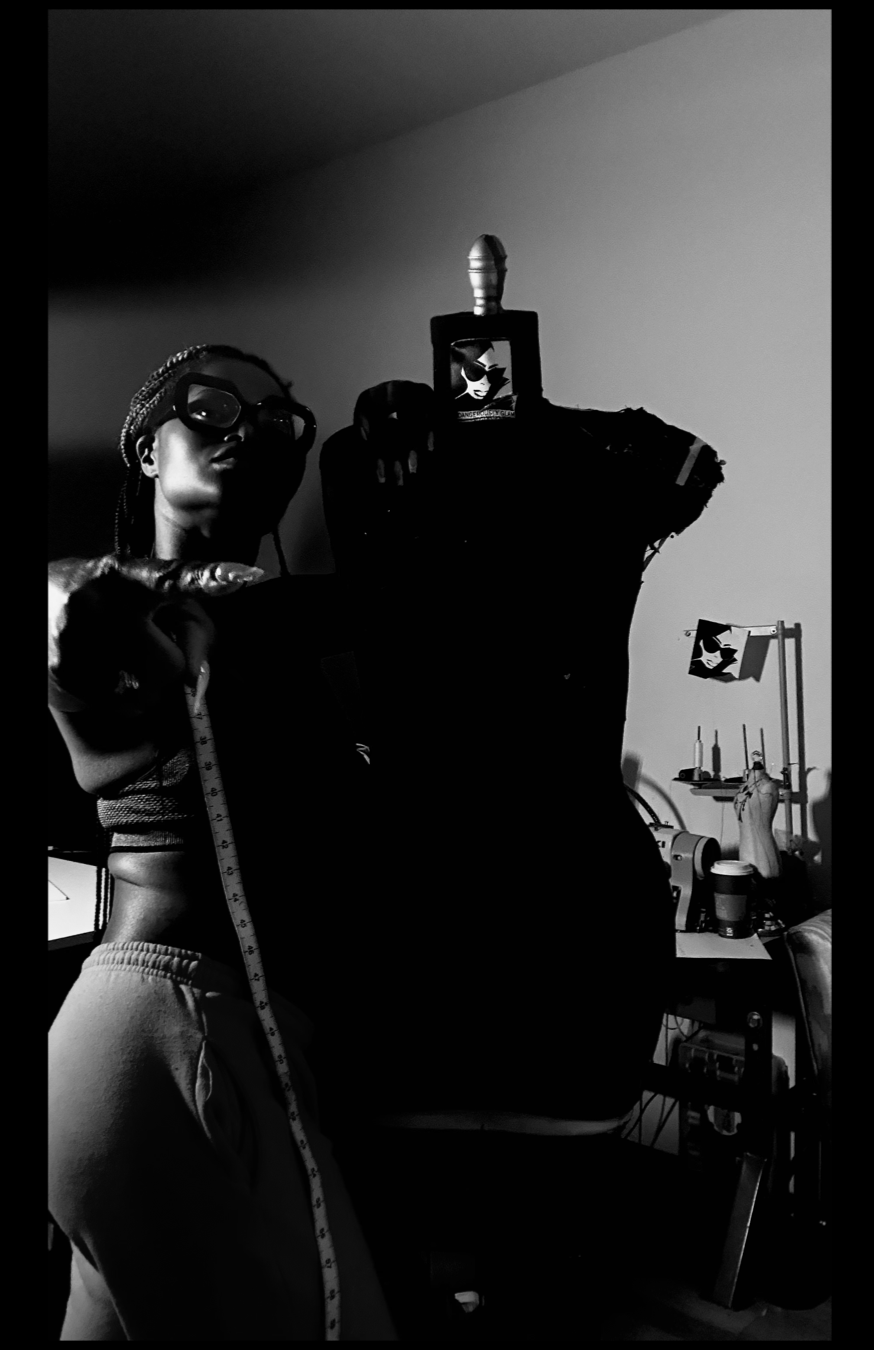 A woman with braided hair and glasses measuring a dress form in a sewing studio. The dress form is draped with a fabric, and there are sewing tools and equipment on a table in the background. The image is in black and white.