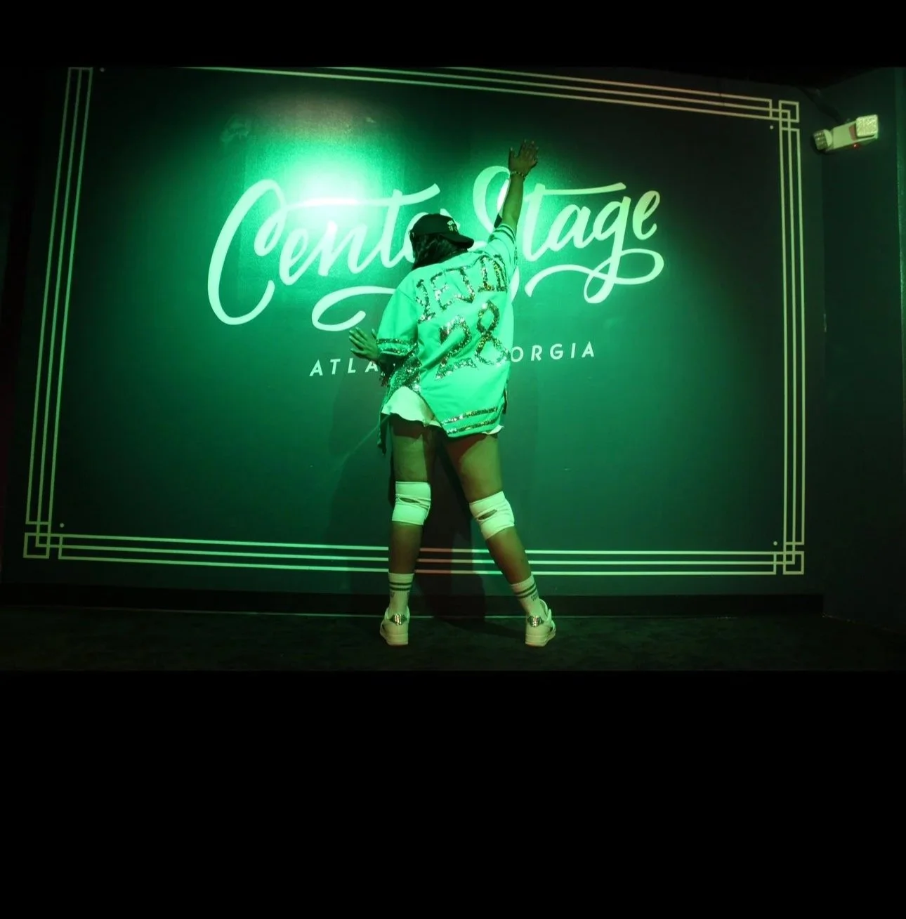 Person standing with back to the camera, raised arm, in front of a neon sign that says 'Center Stage' in lights, Atlanta Georgia, wearing a sports jersey, shorts, sneakers, and knee-high socks, illuminated in green light.