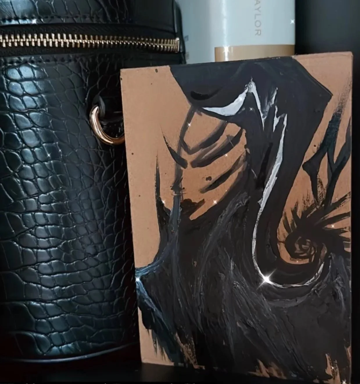 Abstract black, white, and brown painting leaning against a black textured handbag with a gold zipper and ring, with a white and gold container in the background.