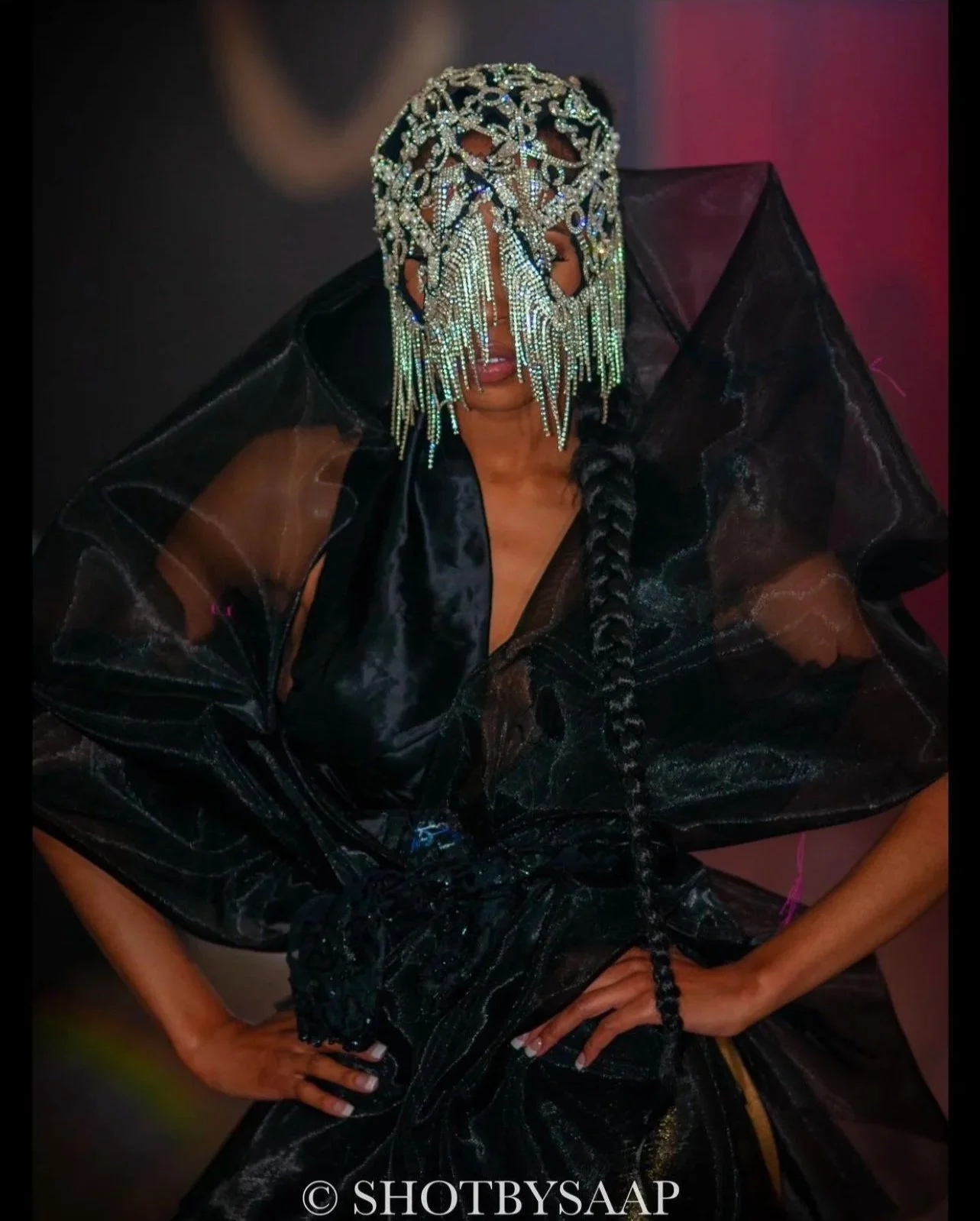 A woman wearing a face cover made of sparkling beads and chains, a sheer black dress with voluminous textured sleeves, and a long braid hairstyle, posing with both hands on her hips.