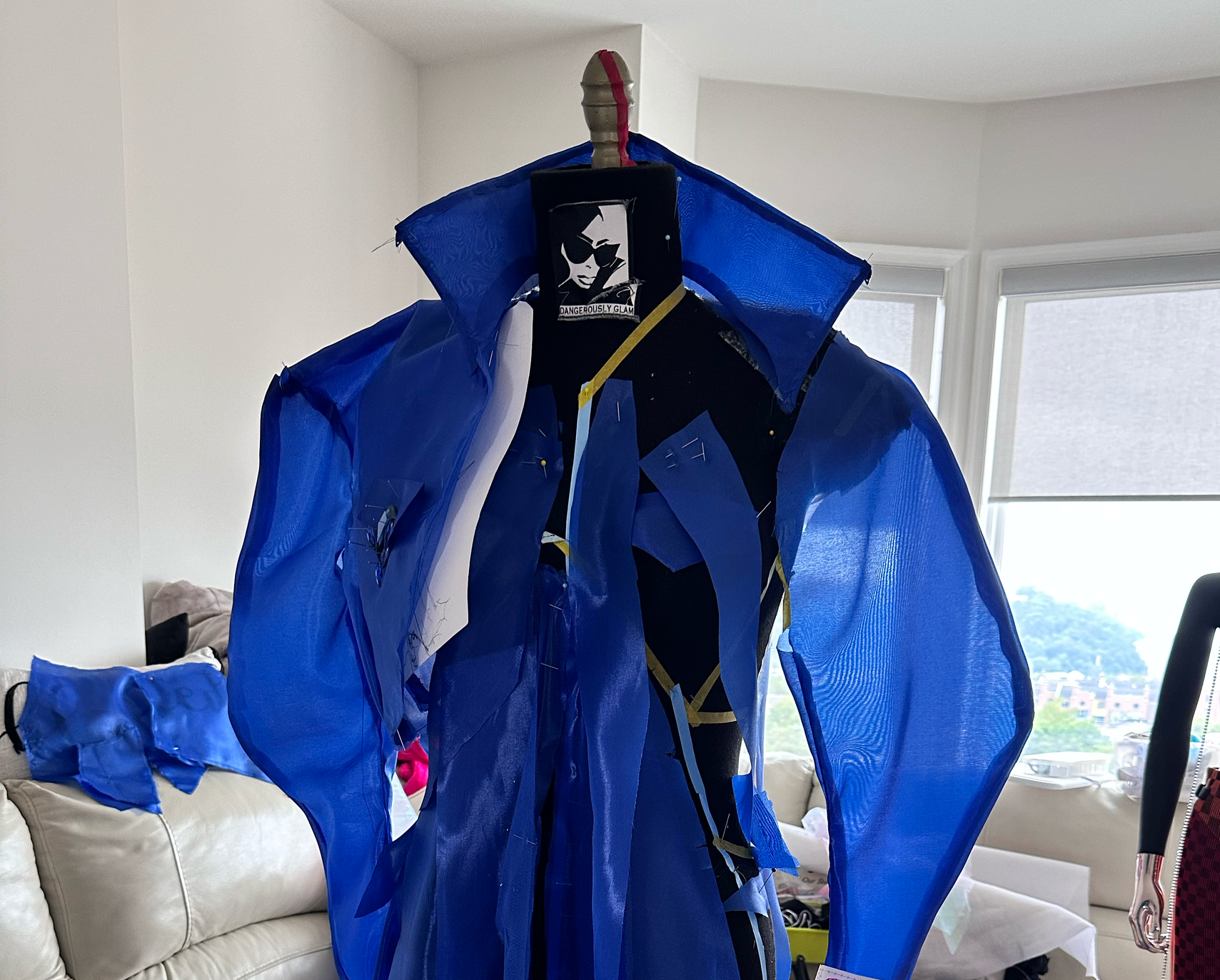 A dress form dressed in a unique, avant-garde blue and black garment with structured, flowing fabric, in a room with large windows and a white couch in the background.