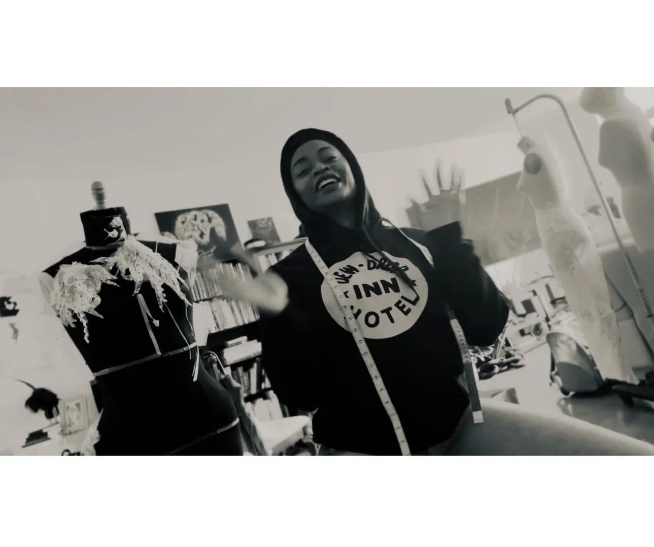 A woman wearing a hoodie and measuring tape around her neck, smiling and gesturing in a room with mannequins and medical or sewing equipment.