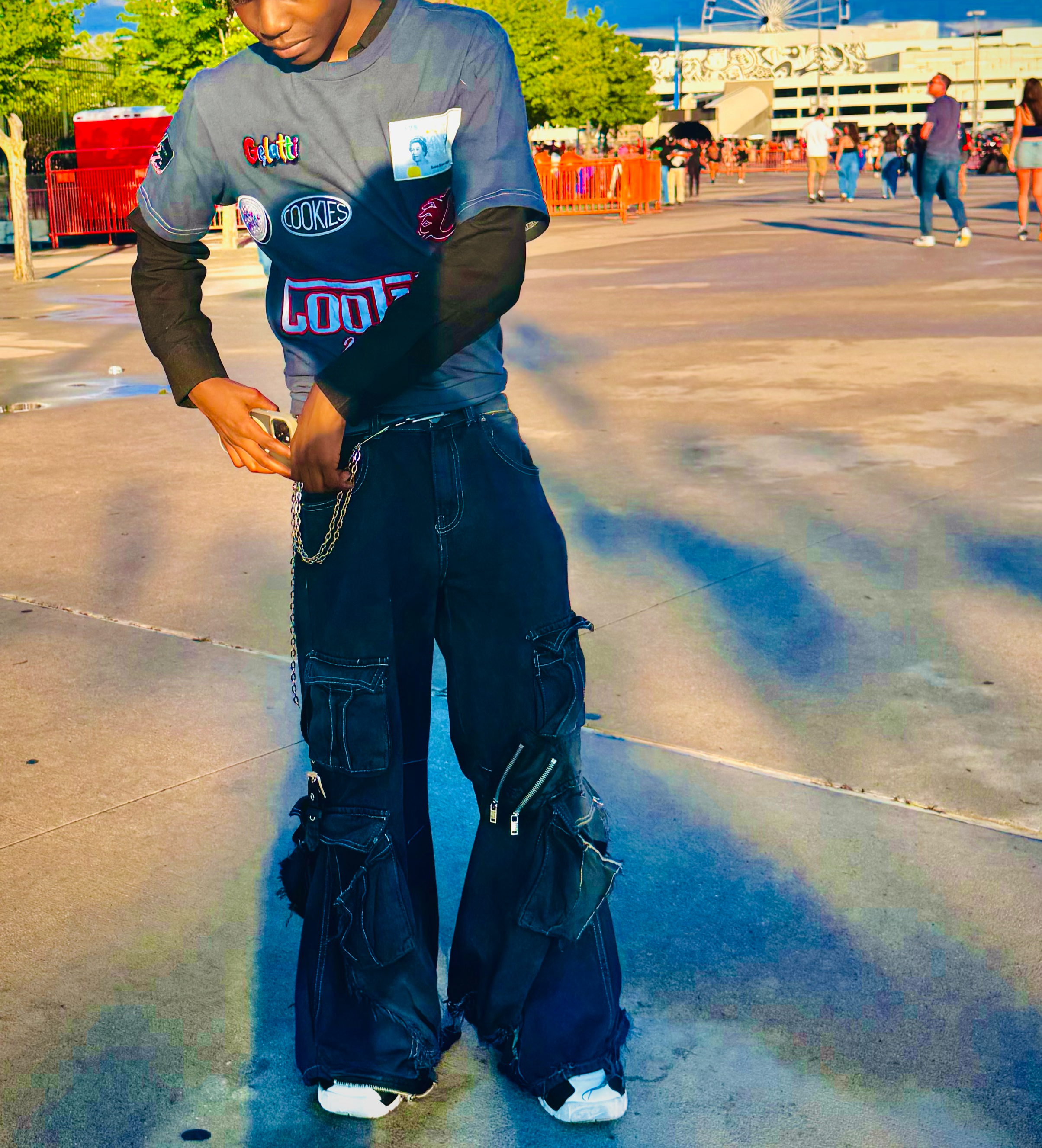 A young person with curly black hair and dark sunglasses wearing a multicolored shirt layered over a long-sleeve black shirt, dark cargo pants with zippers and chains, standing on a paved area at an amusement park. In the background, there are people