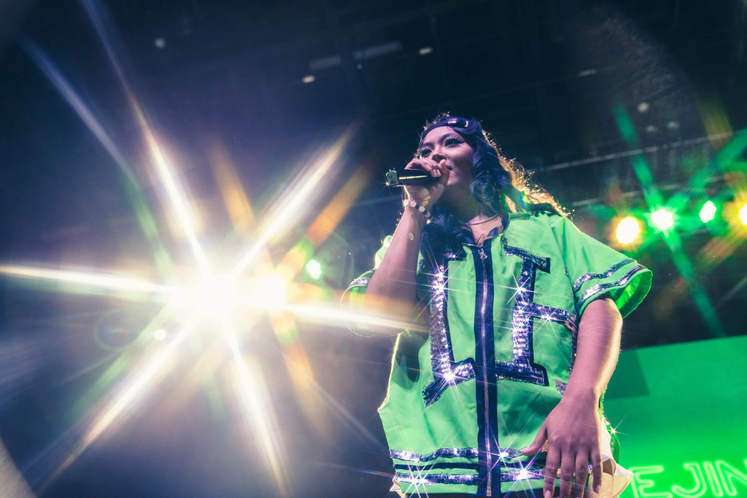 Female singer performing on stage with bright lights and green background, wearing a bright green, sparkly outfit.