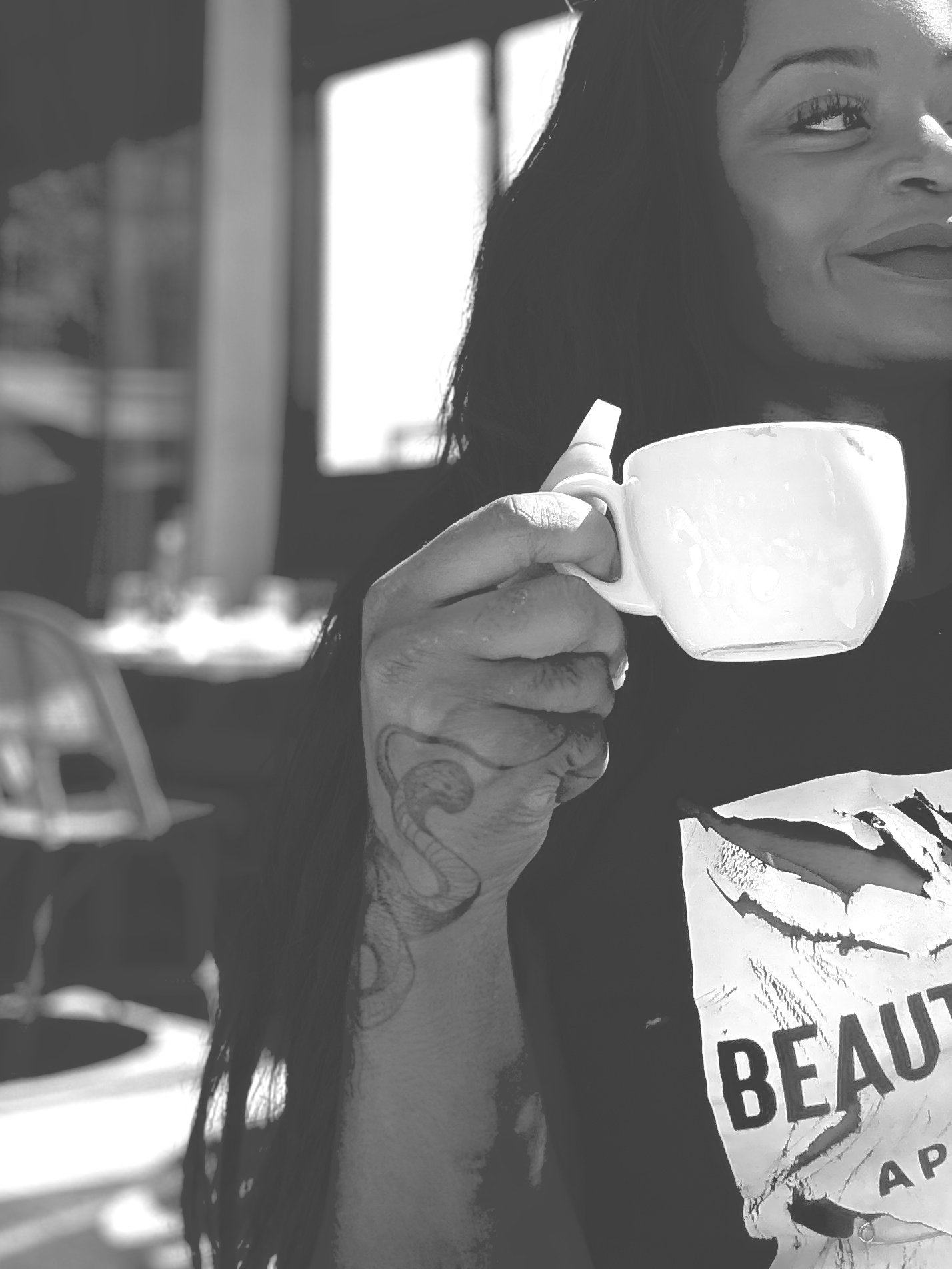 Woman holding a white mug, smiling, in a casual setting with sunlight streaming in.