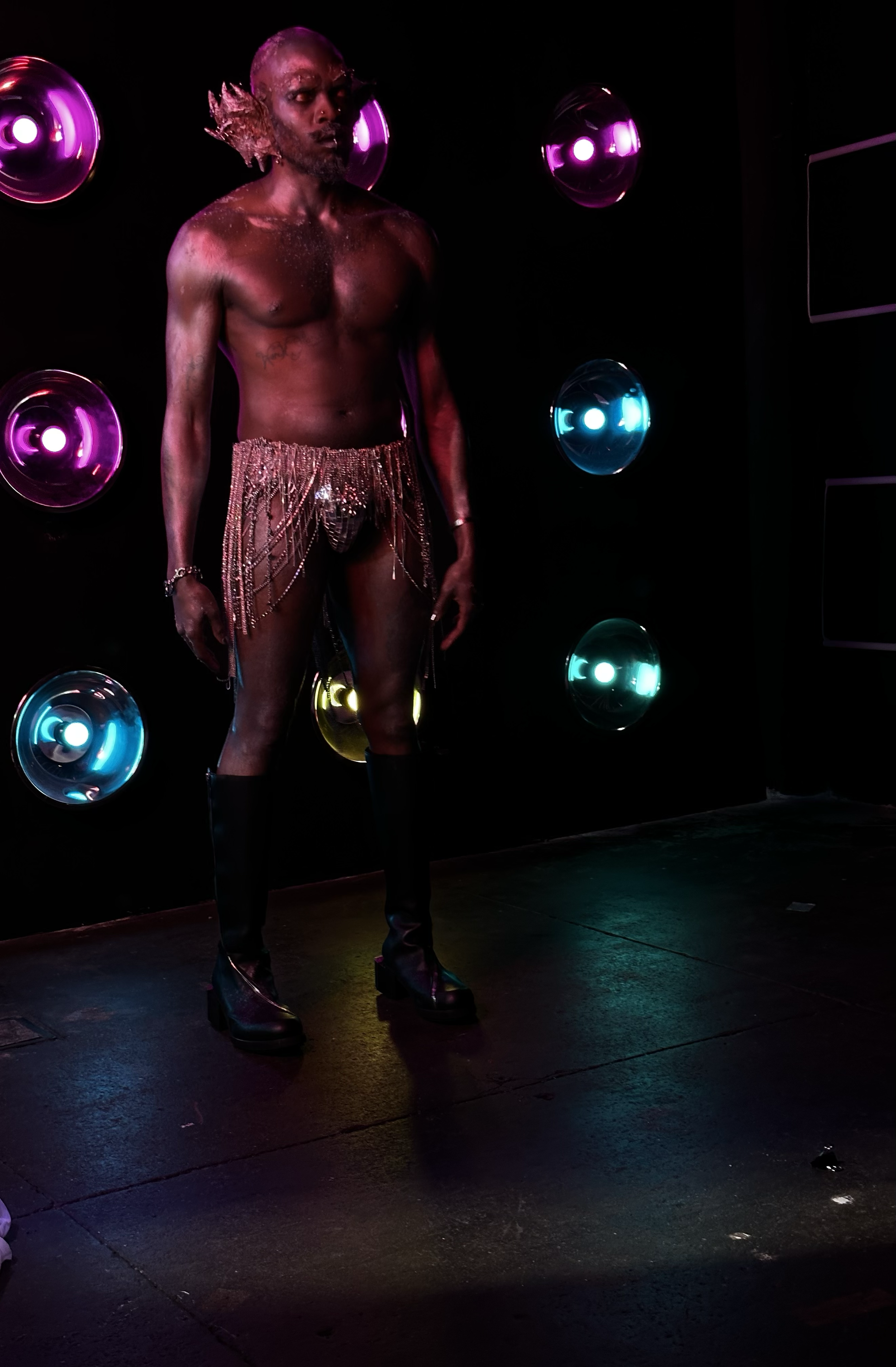 A person standing on a dark stage with colorful lighting, wearing dramatic makeup and elaborate clothing with a beaded skirt and over-the-knee boots.