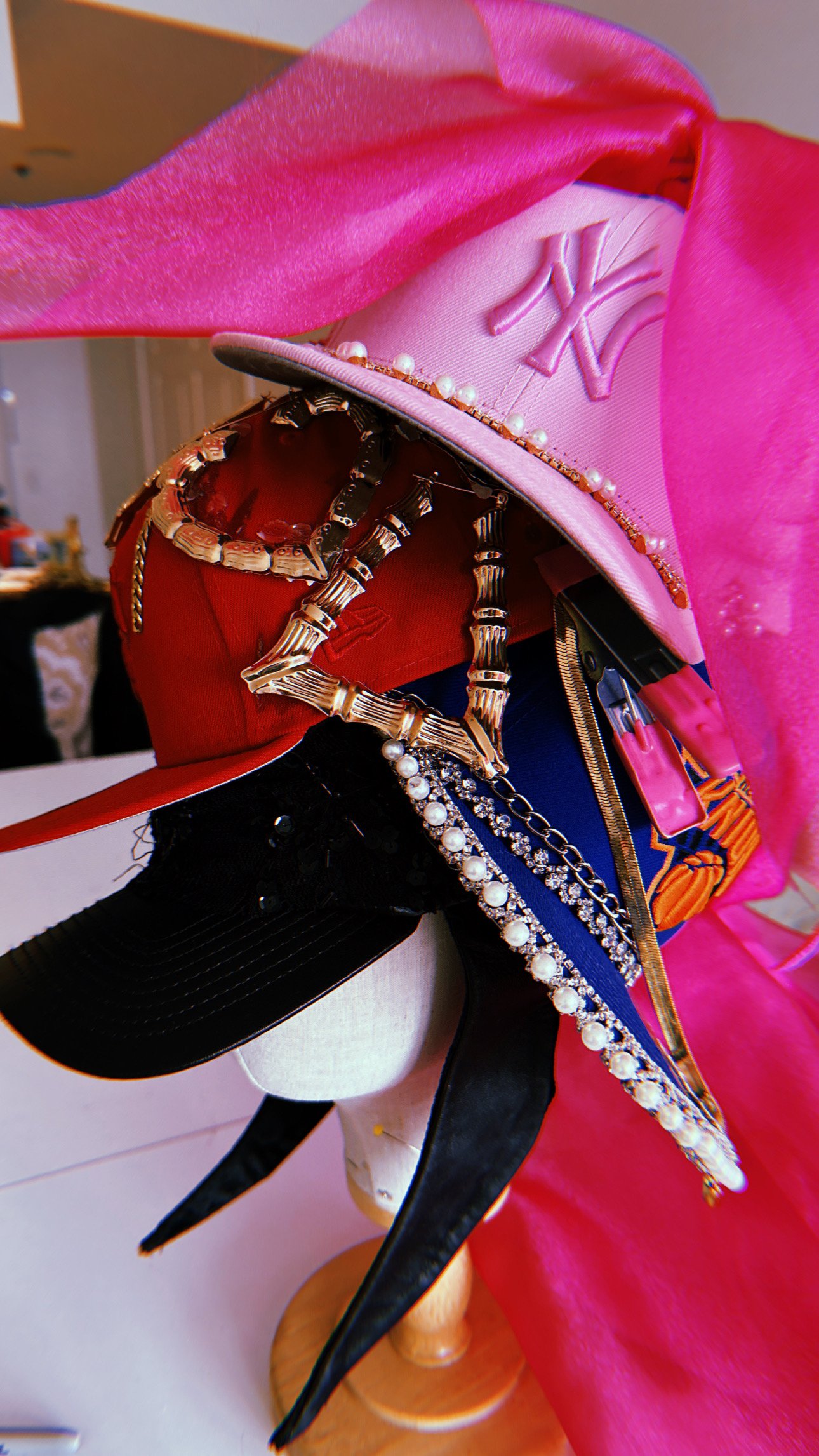 A display of various fashion accessories including a pink cap with the New York Yankees logo, a black baseball cap, a red baseball cap, a pink scarf, necklaces with pearls and rhinestones, some jewelry, and a pink item resembling a handbag or pouch.