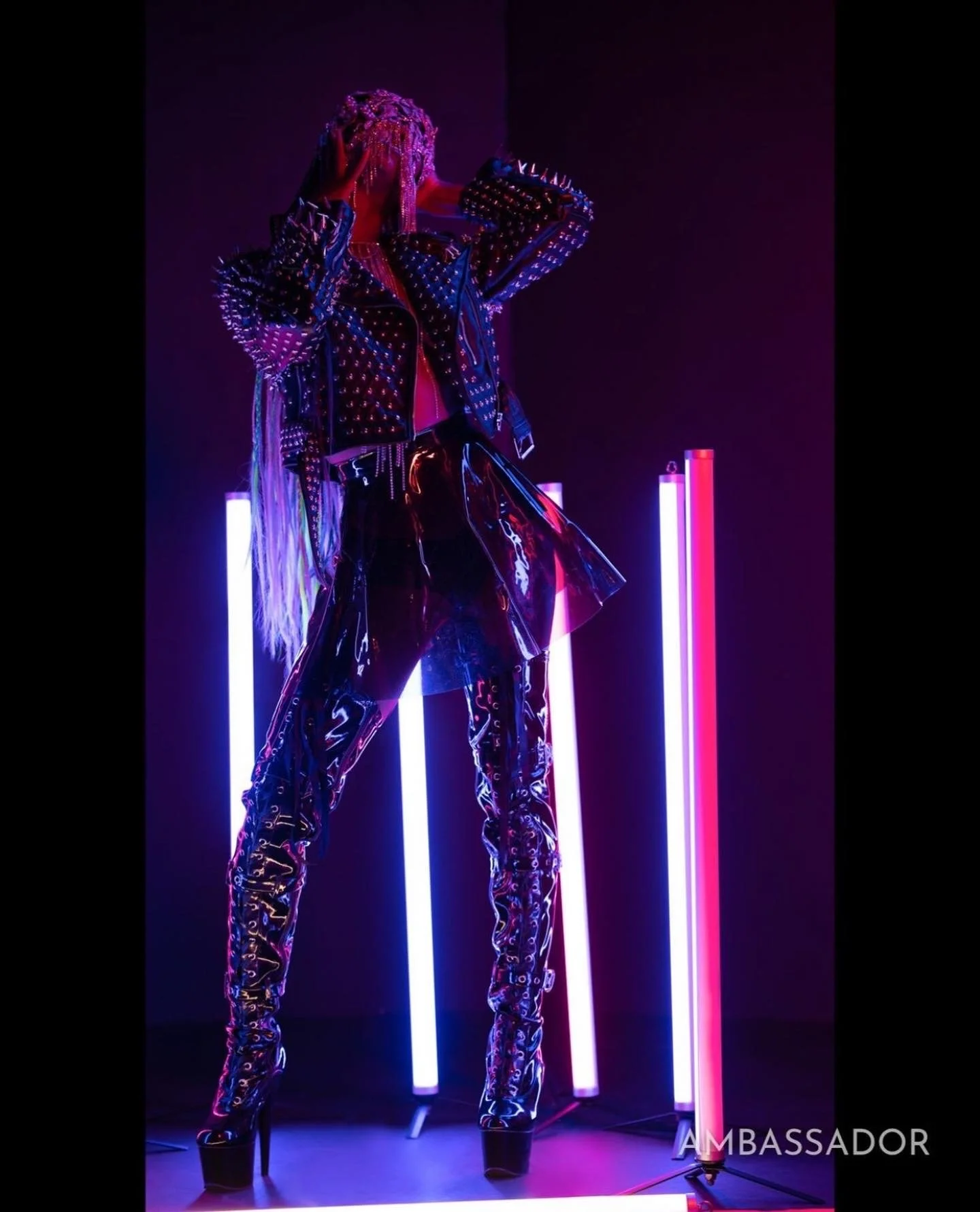 Person dressed in a studded leather jacket and shiny pants, wearing a headpiece, standing in front of colorful neon lights.