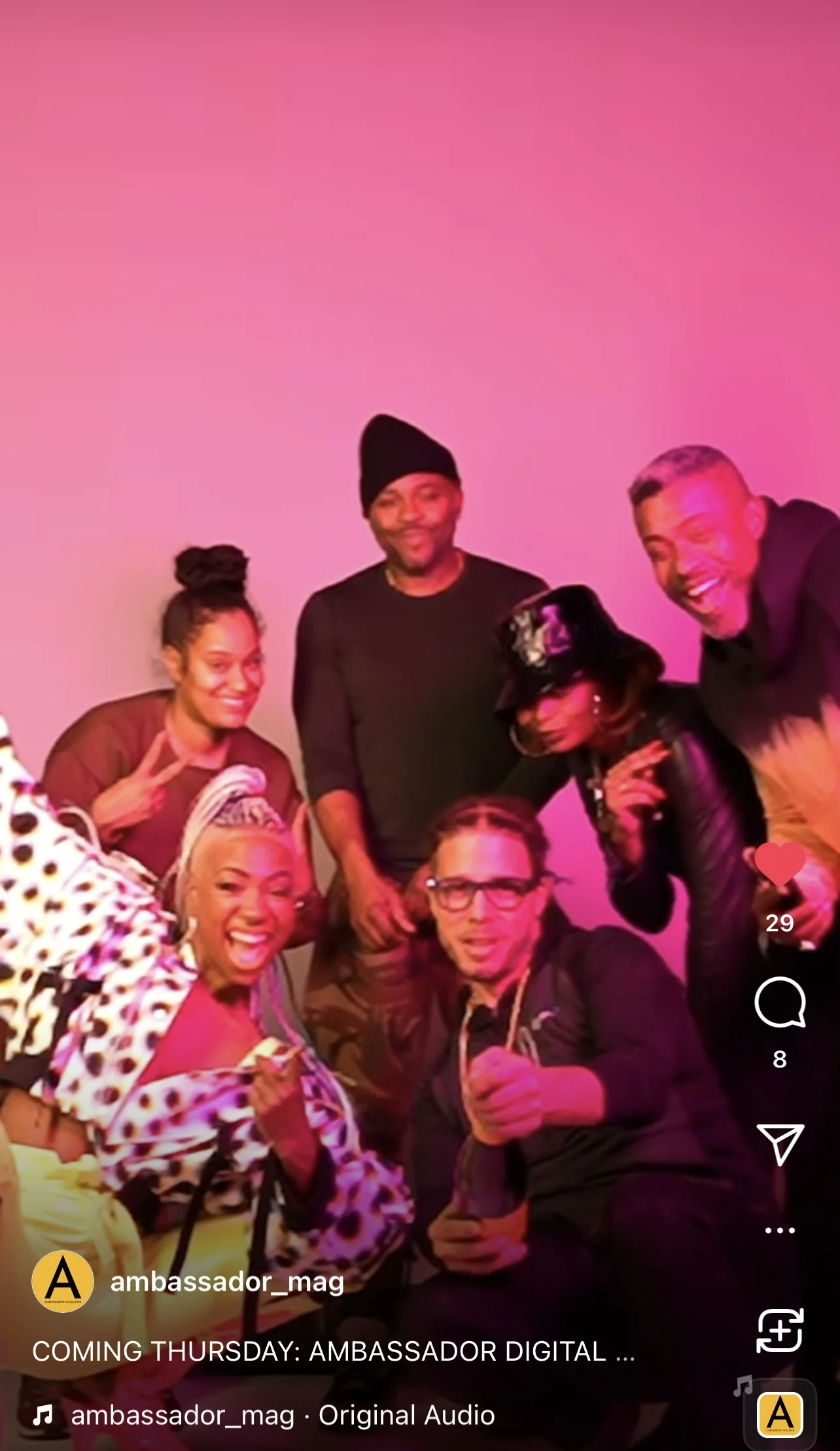 Group of six diverse people, four women and two men, gathered together, smiling and posing for a photo against a pink background. Some are making peace signs, and one person is holding a drink.