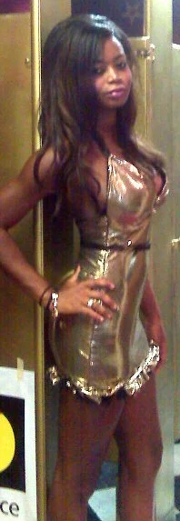 Young woman with long wavy hair wearing a shiny, gold-colored dress in front of a mirror in a room with gold-colored cabinets.