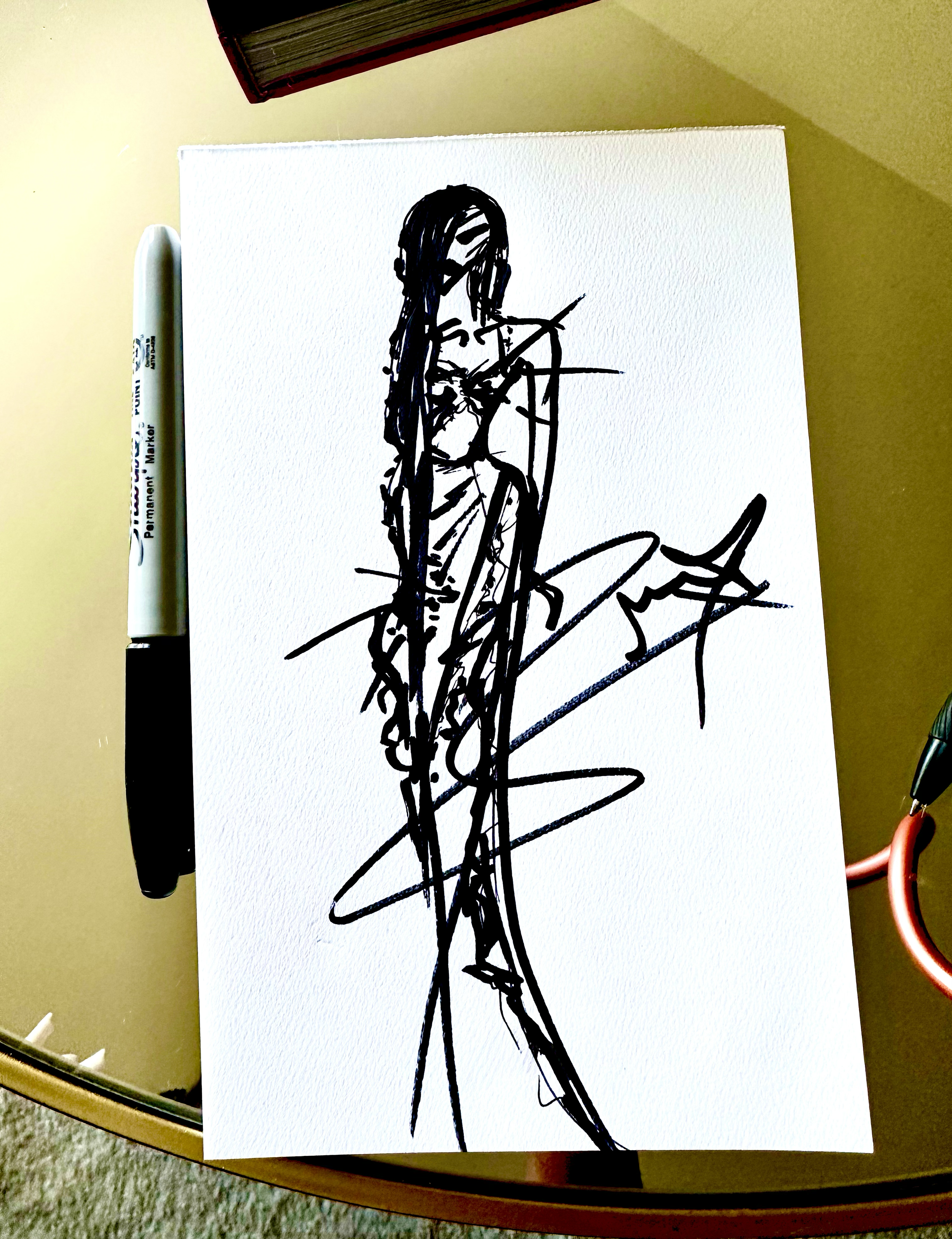 A black ink sketch of a woman with long hair, sitting with one leg crossed over the other, on white textured paper. The sketch is minimalistic and abstract, with bold, loose lines.