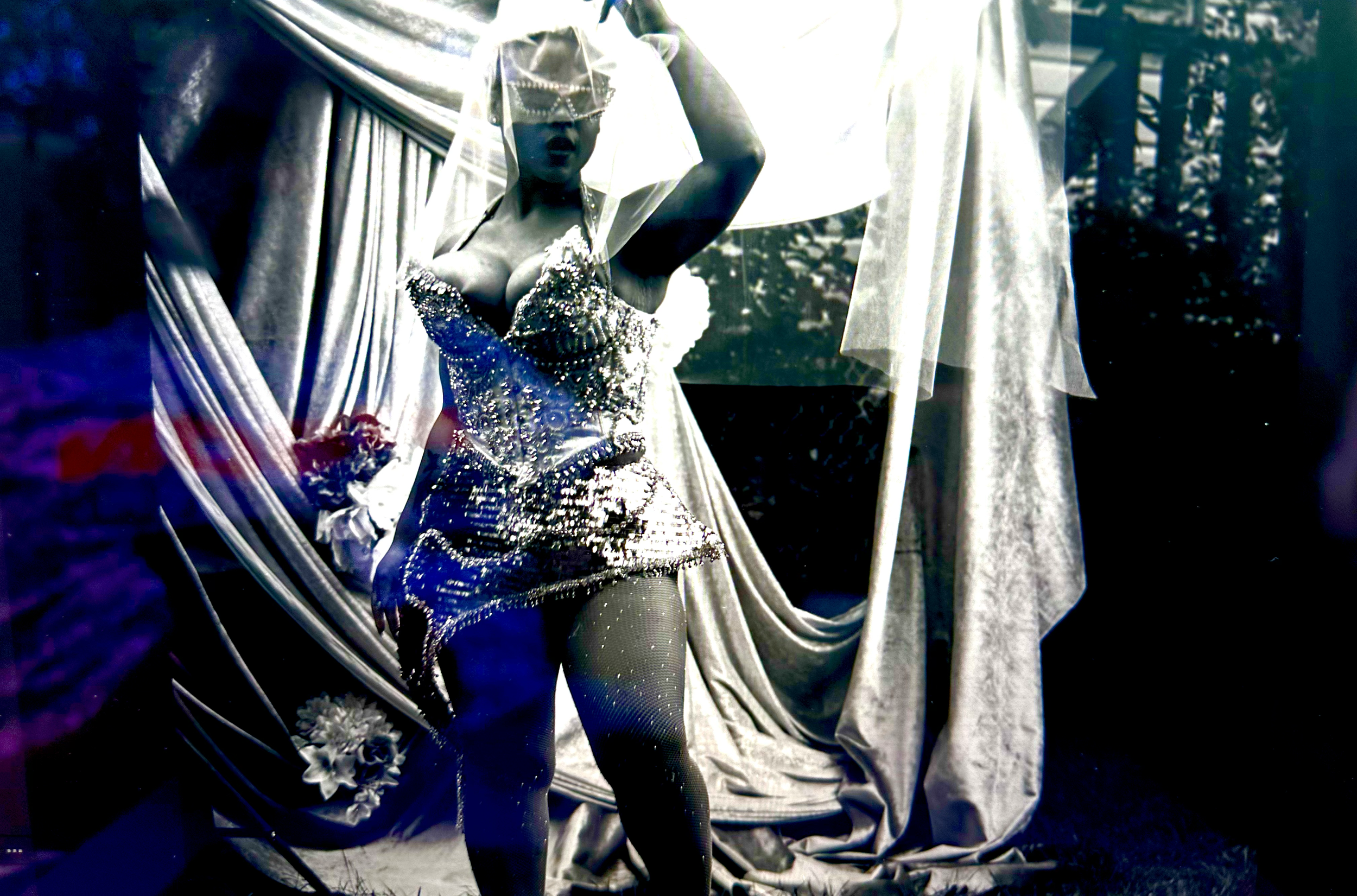 A person dressed in a shiny, sequined outfit with a matching headpiece and veil, standing outdoors in front of draped fabric with flowers and trees in the background.