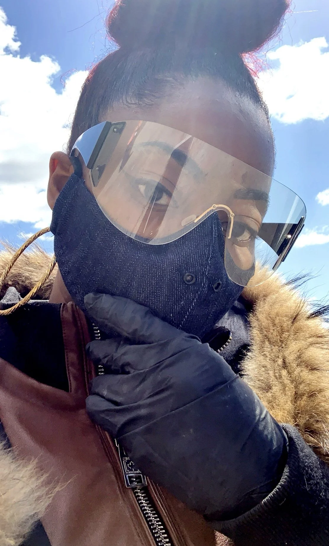 A person wearing sunglasses, a face mask, and a brown jacket with a fur-lined hood, holding their hand near their face against a bright blue sky with clouds.
