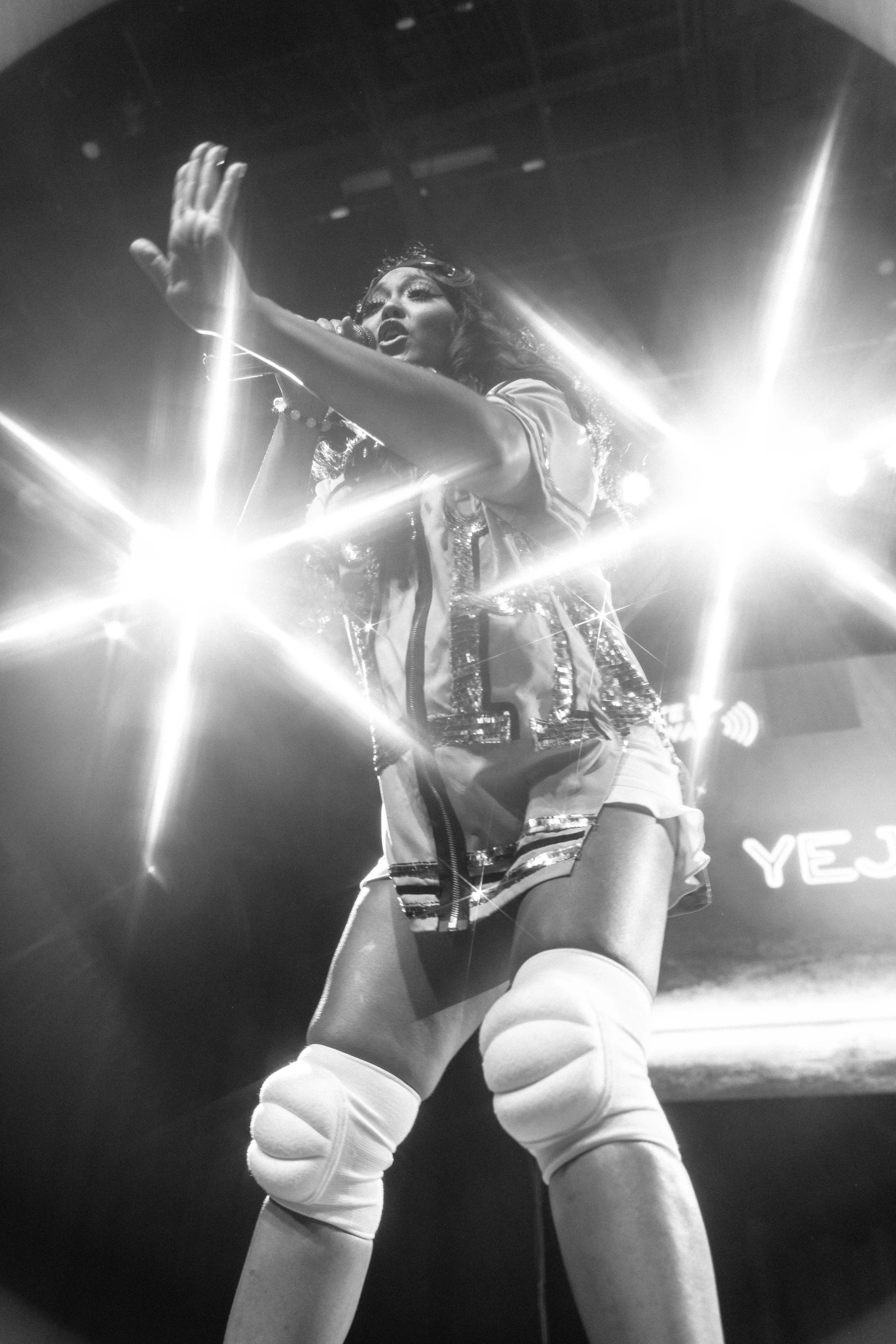 Performer on stage singing into a microphone, wearing a shiny vest, shorts, and knee-high socks, with bright stage lights creating starburst effects.