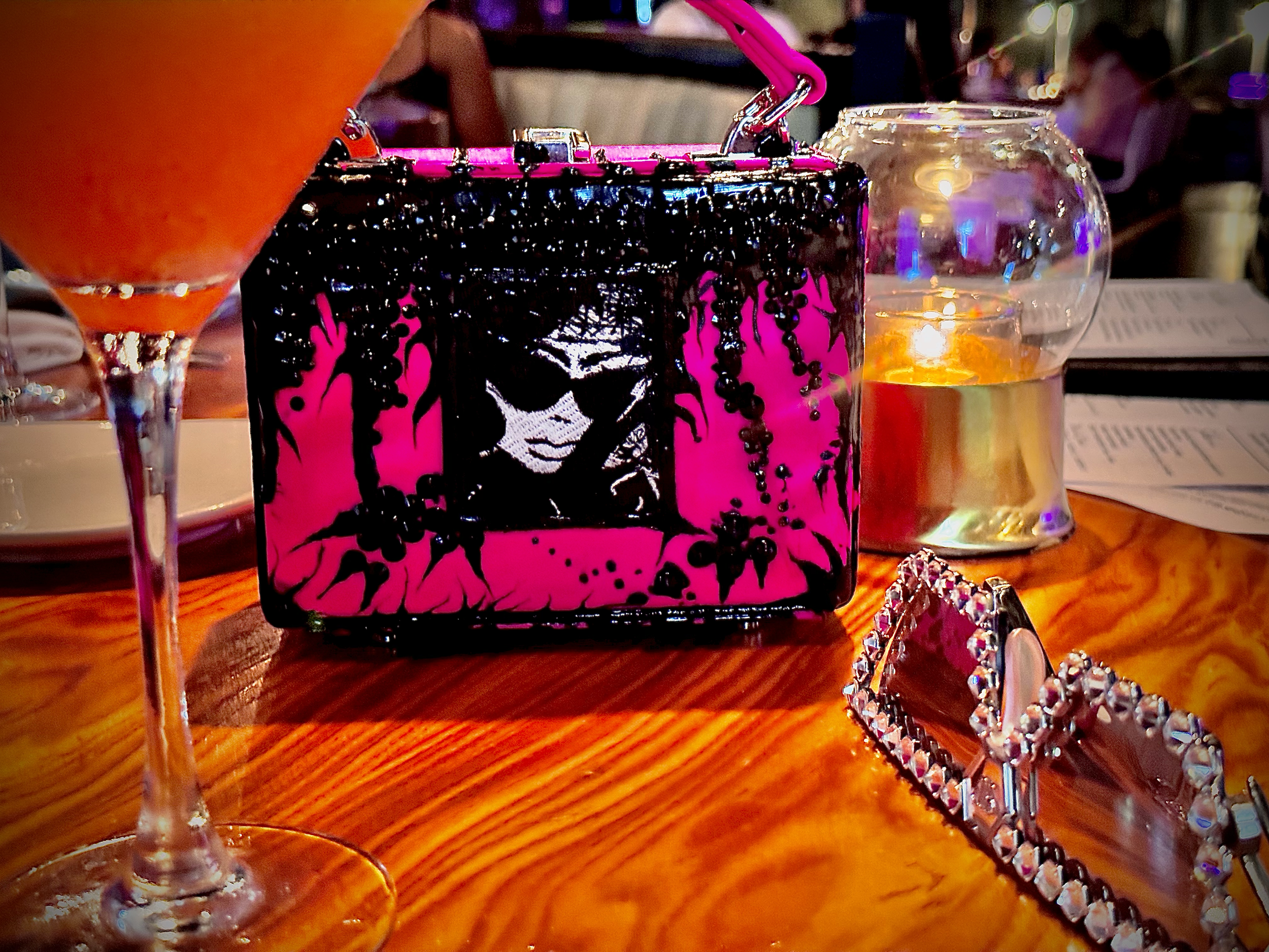 A pink and black handbag with an illustration of a woman and a beaded chain, a glass of orange drink, a small glass jar with a candle, and a jewelry piece with rhinestones on a wooden table at a restaurant or cafe.
