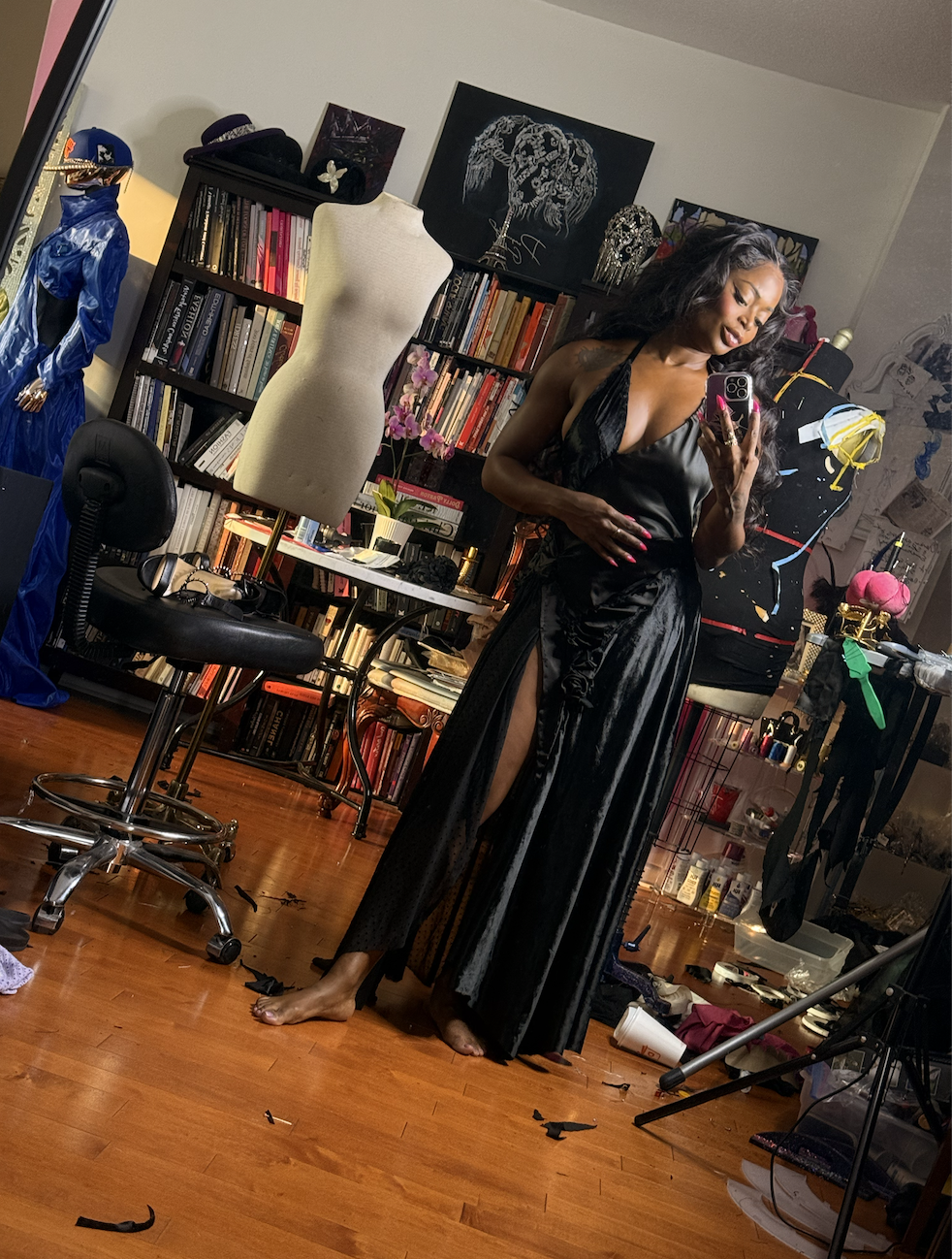 Woman taking a mirror selfie in a room with a bookshelf, dress form, mannequin, and art supplies, wearing a black satin gown with a high slit.
