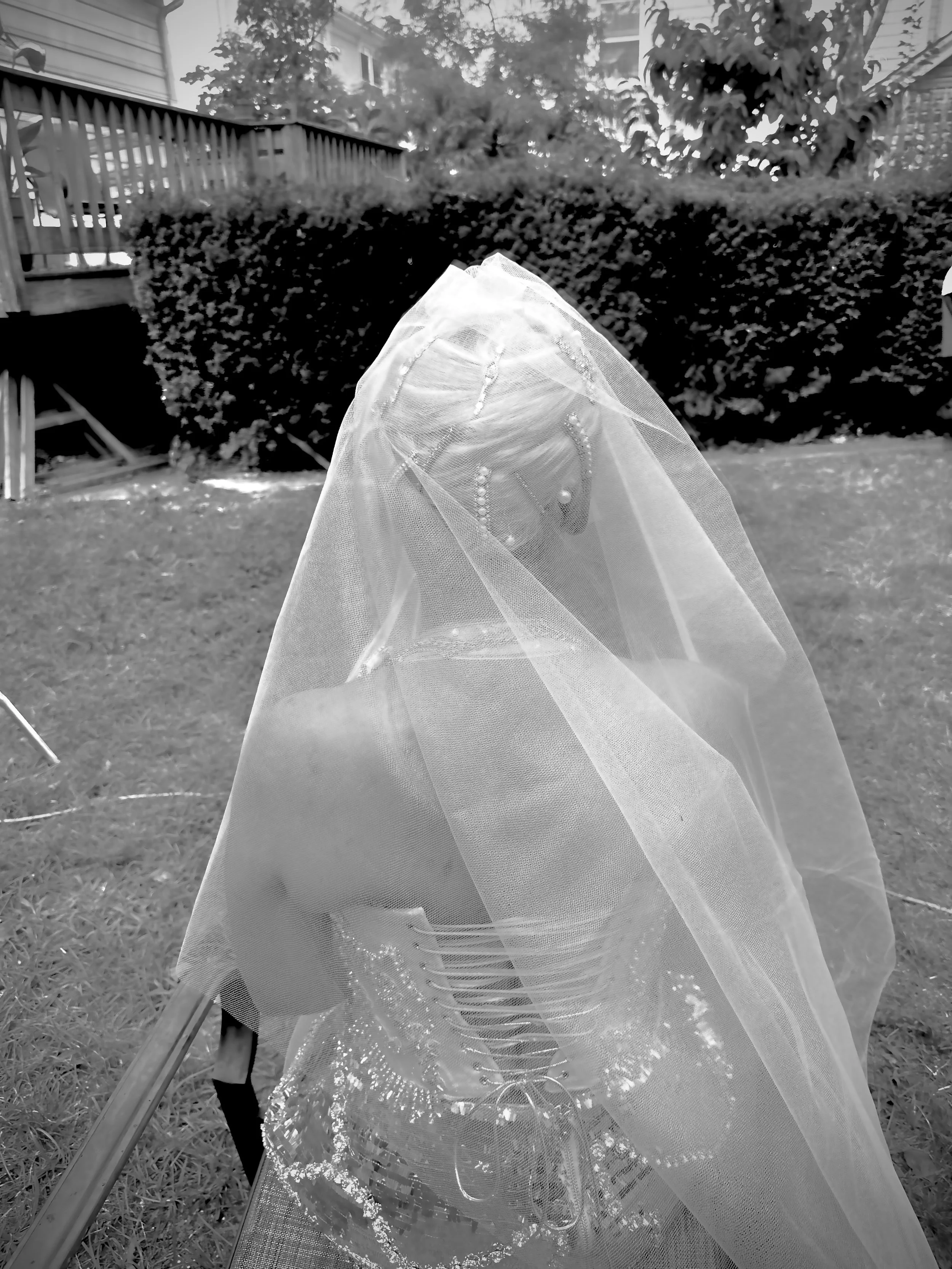 A skeleton adorned with jewelry and dressed in a veil, sitting outdoors on a chair in a backyard with a hedge and trees in the background.