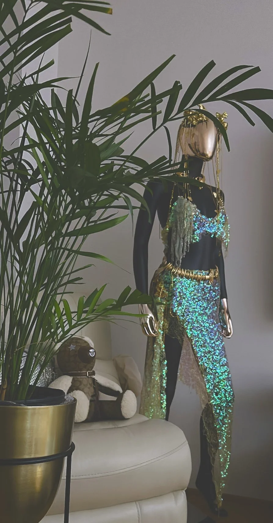 A black mannequin with a shiny metallic gold headpiece and various layered gold accessories, dressed in a sparkling, holographic, green and gold outfit, standing near a white sofa with a stuffed teddy bear and a potted plant in the foreground.