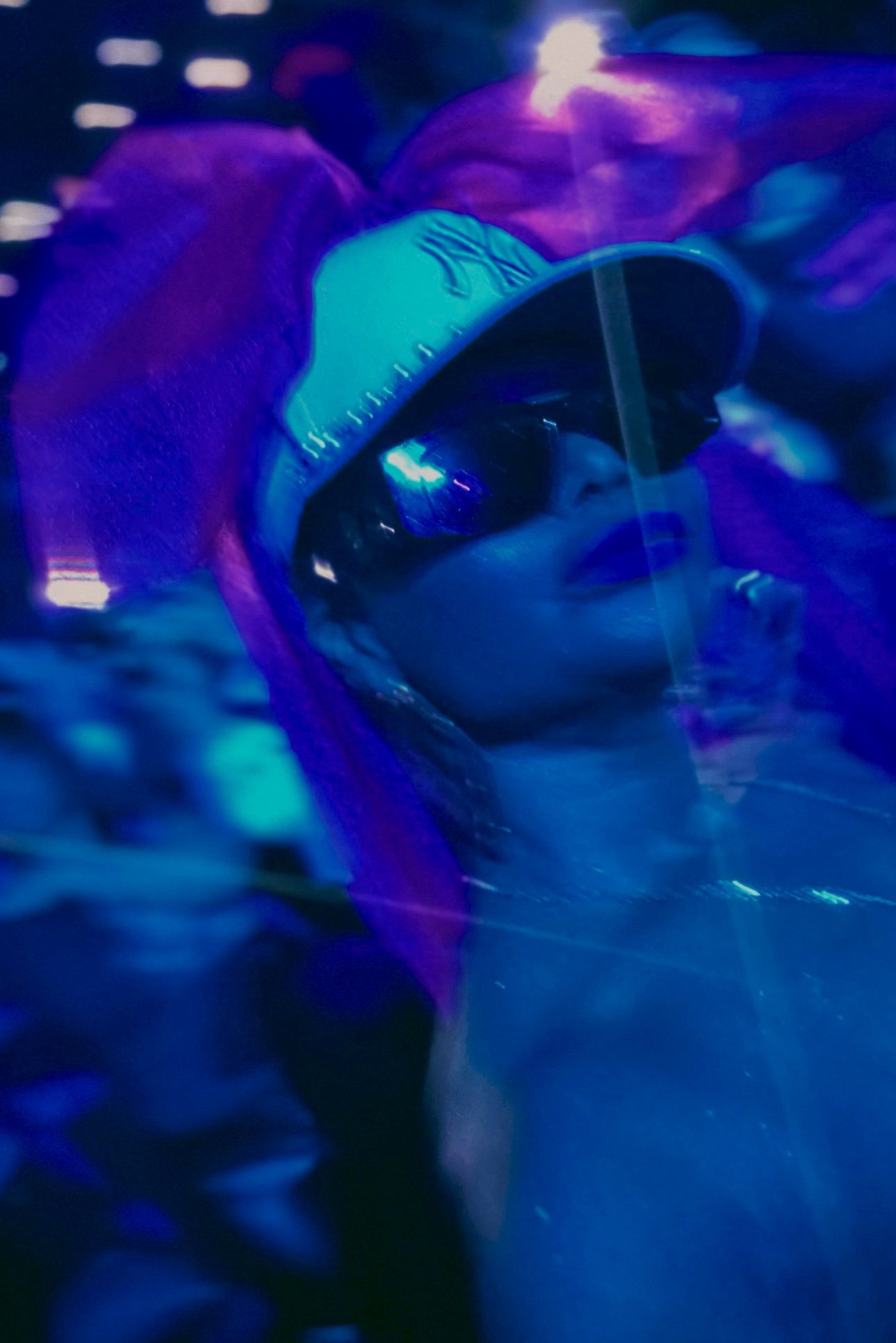 Person wearing sunglasses, a baseball cap, and a pink wig in a dark, neon-lit environment.