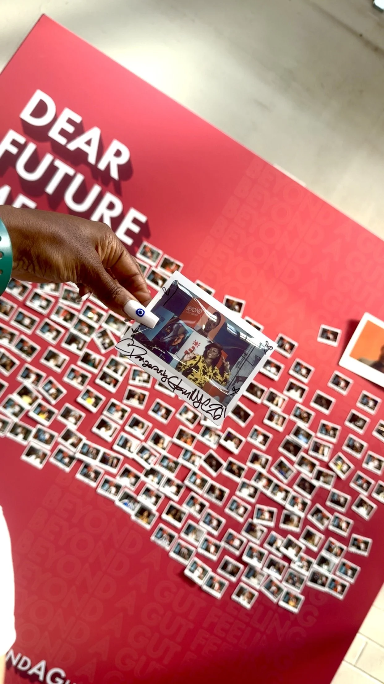 Close-up of a person's hand holding a signed photo in front of a letter board displaying many small printed photographs, with a red backdrop that reads 'Dear Future' in large white letters.