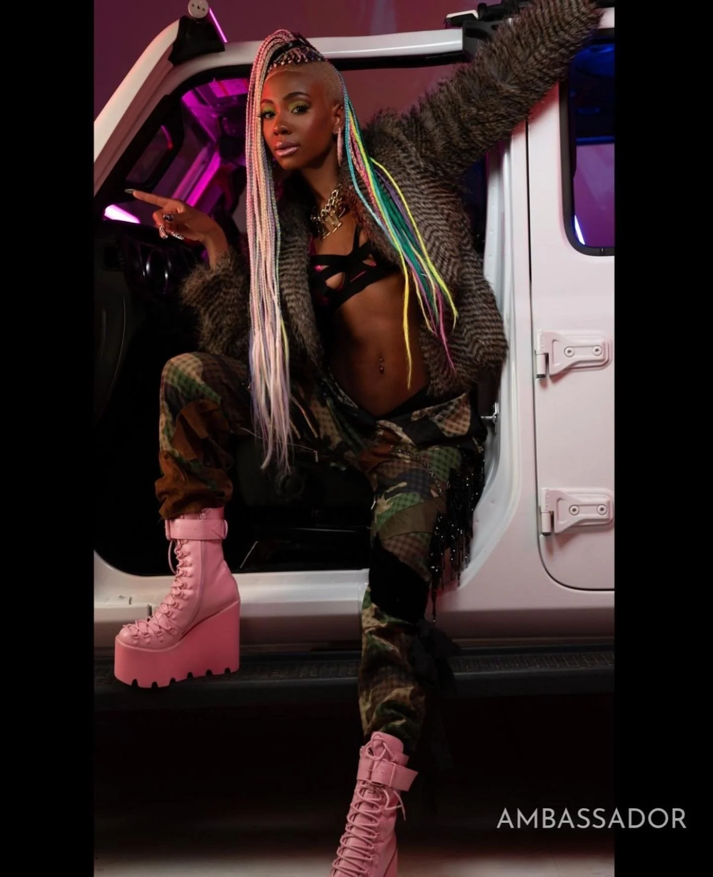 A woman with colorful braided hair, wearing a fur coat, camouflage pants, and pink platform boots, sitting on a car's door frame with neon pink lighting in the background.