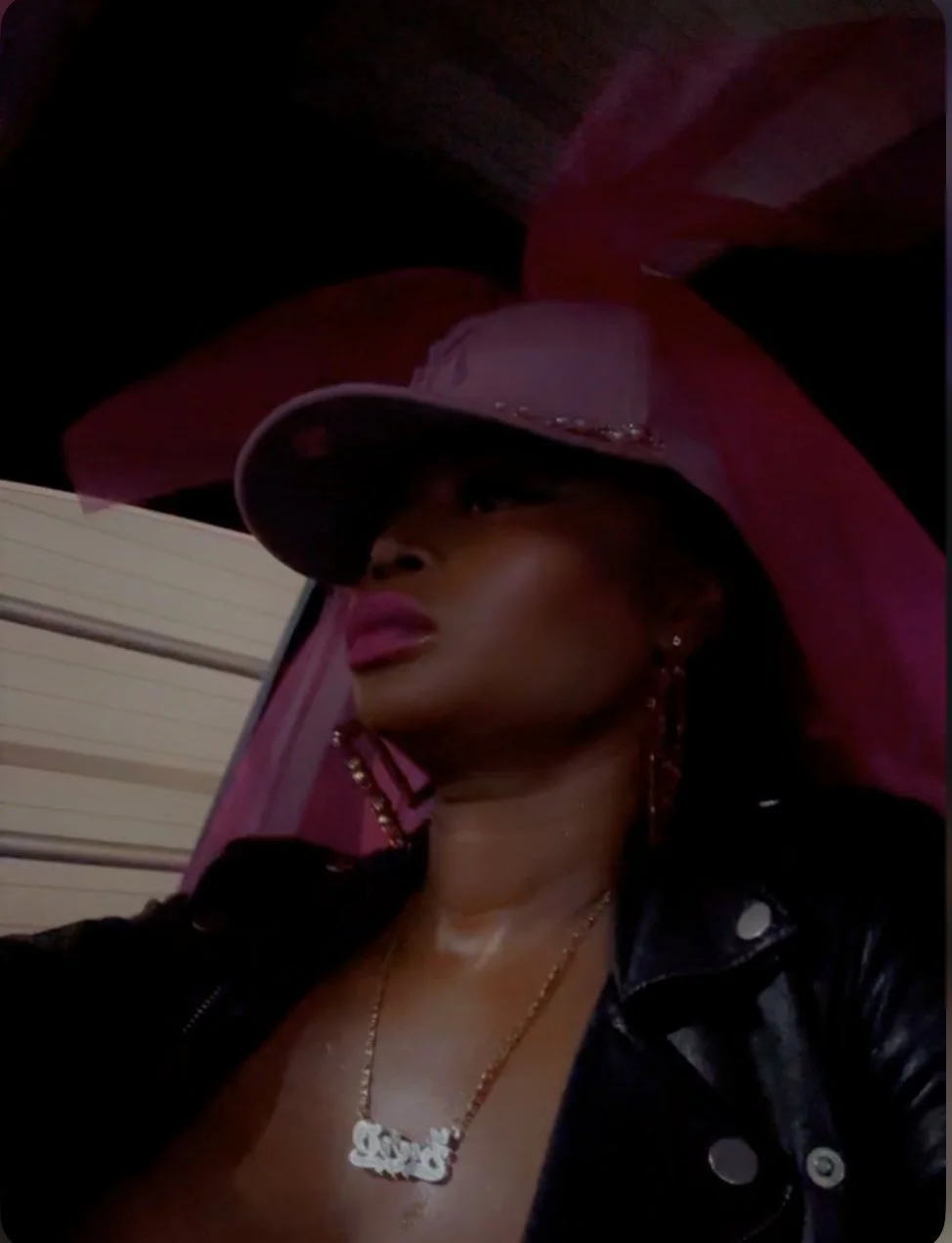 A woman wearing a large pink hat, black leather jacket, and a gold necklace with a nameplate, posing in a dimly lit setting.