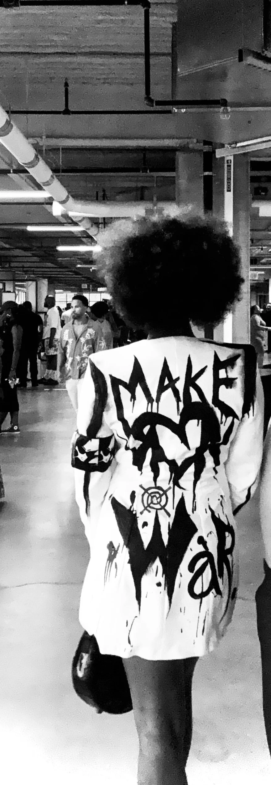 A person with an afro hairstyle walking in an indoor, industrial-style space filled with people. They are wearing a white jacket with black graffiti-style writing and designs on the back.
