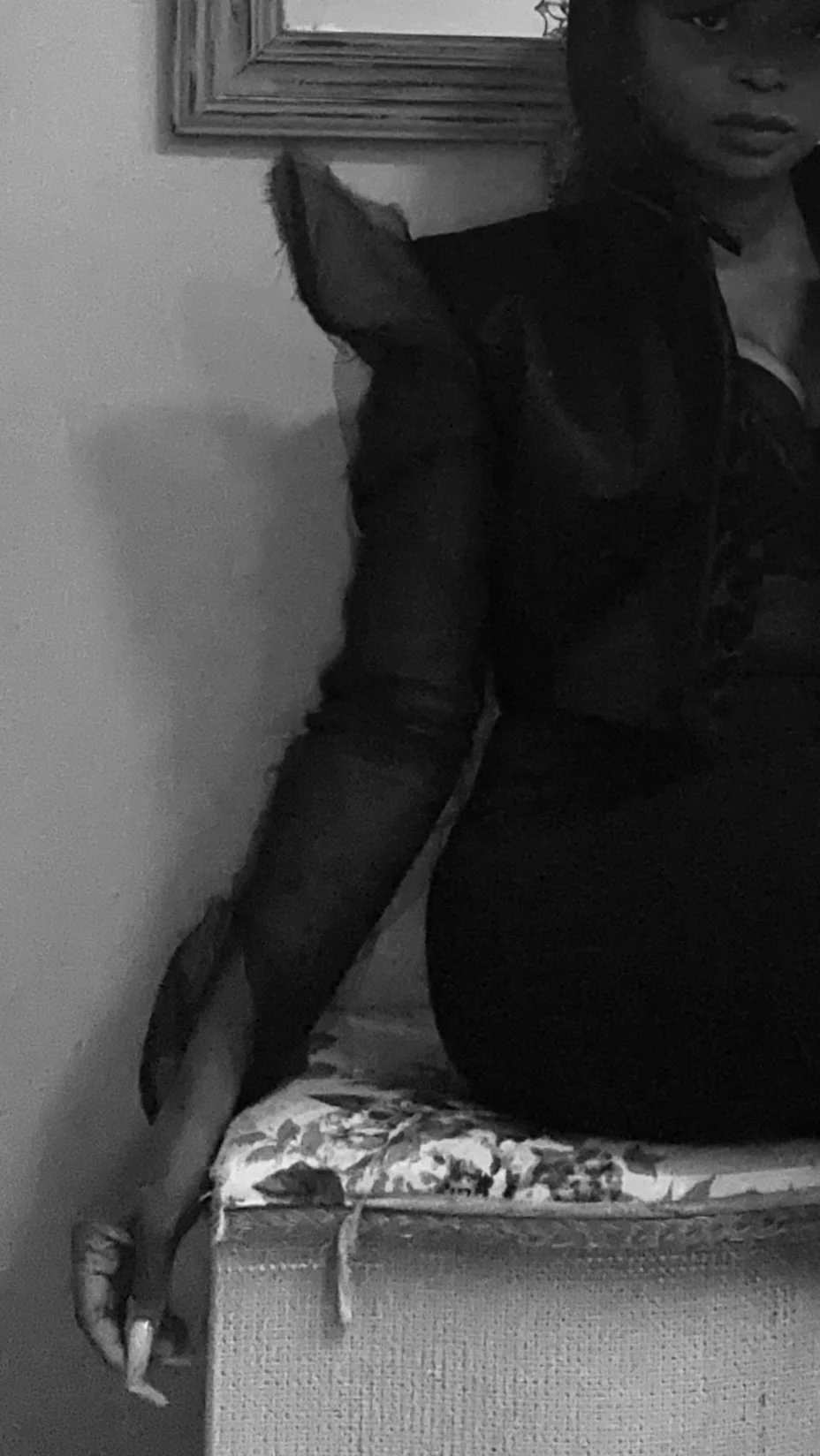 A woman sitting on a floral cushion with her body turned away and her head turned towards the camera, displaying a serious expression. She is wearing a black jacket and has a dog sitting next to her, partially out of the frame, with an attentive post