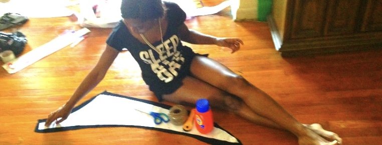 A girl sitting on a wooden floor working on a sewing or craft project, with scissors and tape on paper in front of her.