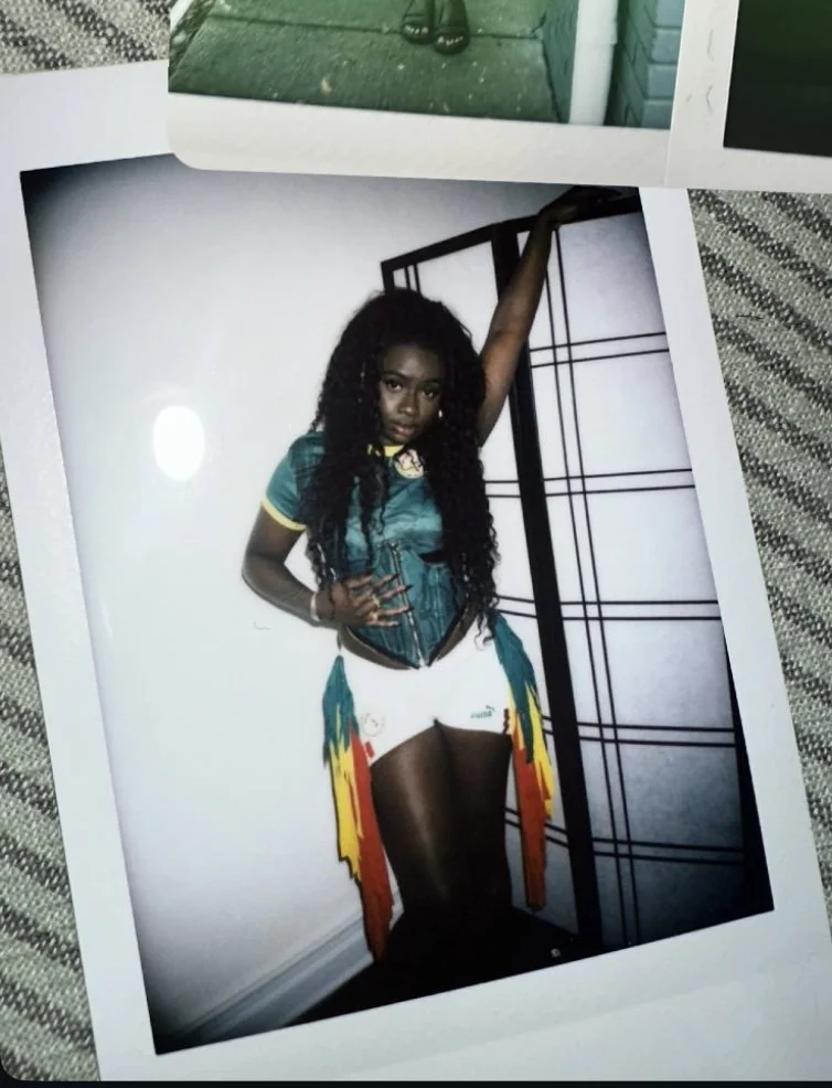 Photo of a woman with long curly hair wearing a teal and yellow top, white shorts, and colorful feathered accessories, standing against a white wall with a black room divider.