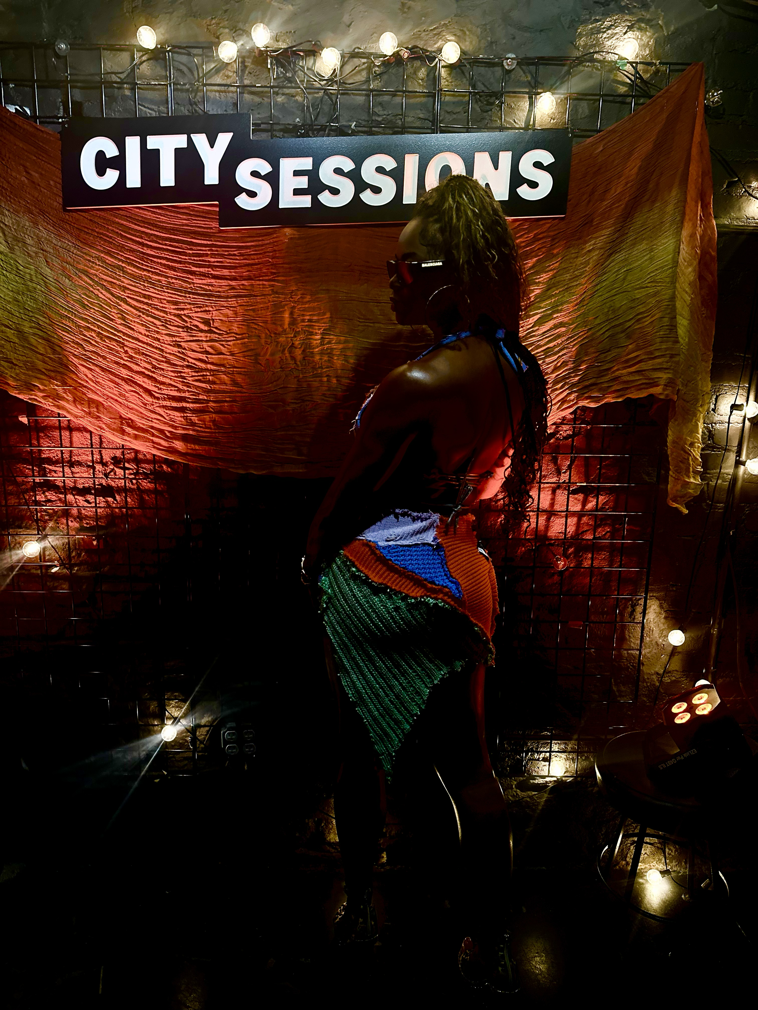 A woman dressed in a colorful, sequined skirt with a sheer top and sunglasses, standing in front of a neon-lit sign that reads 'CITY SESSIONS' in a dimly lit, industrial-style venue with string lights and a textured wall.