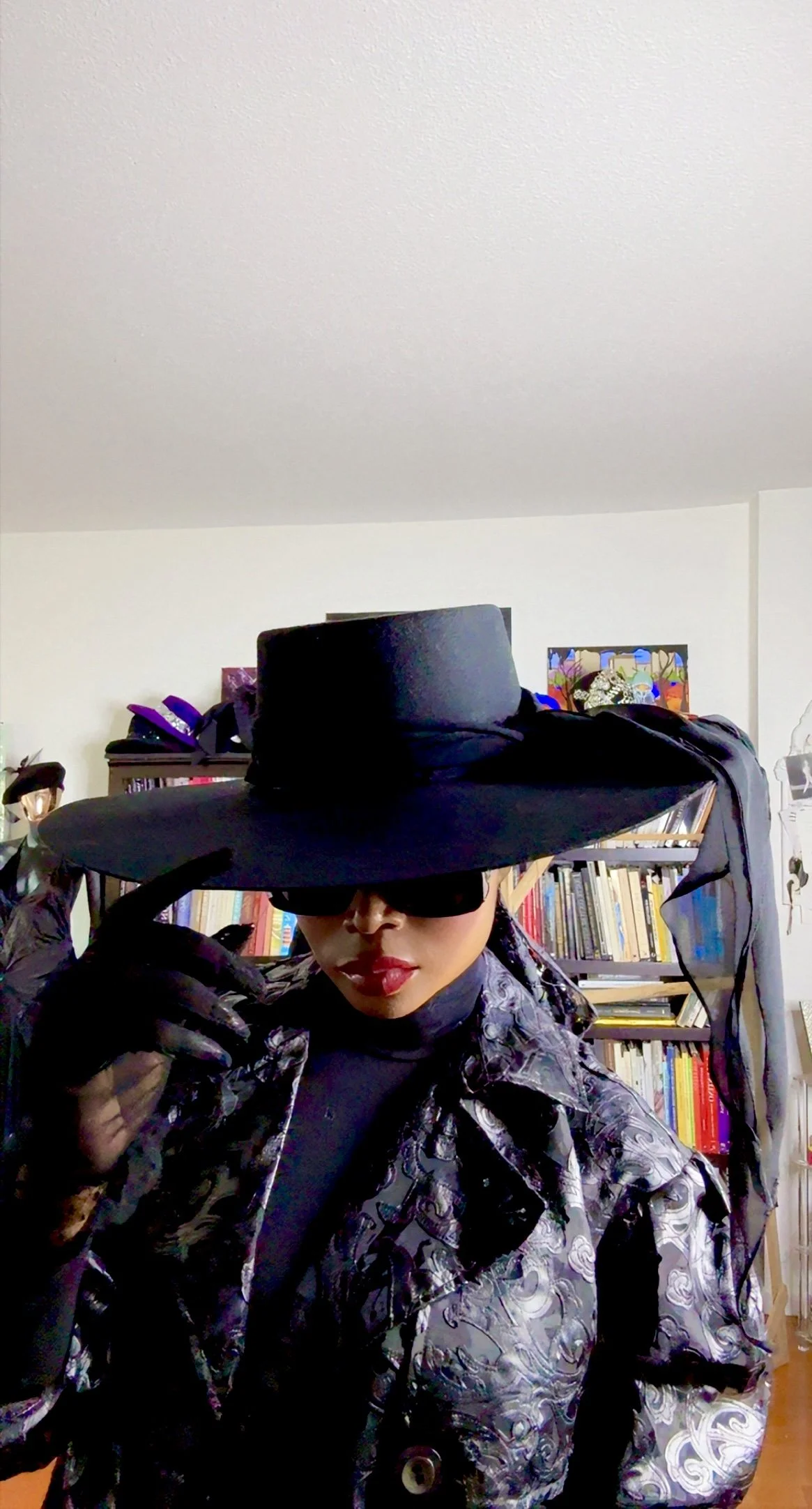 Person wearing a large black hat with a wide brim, black sunglasses, a patterned black and white jacket, and black gloves, standing indoors with bookshelves in the background.