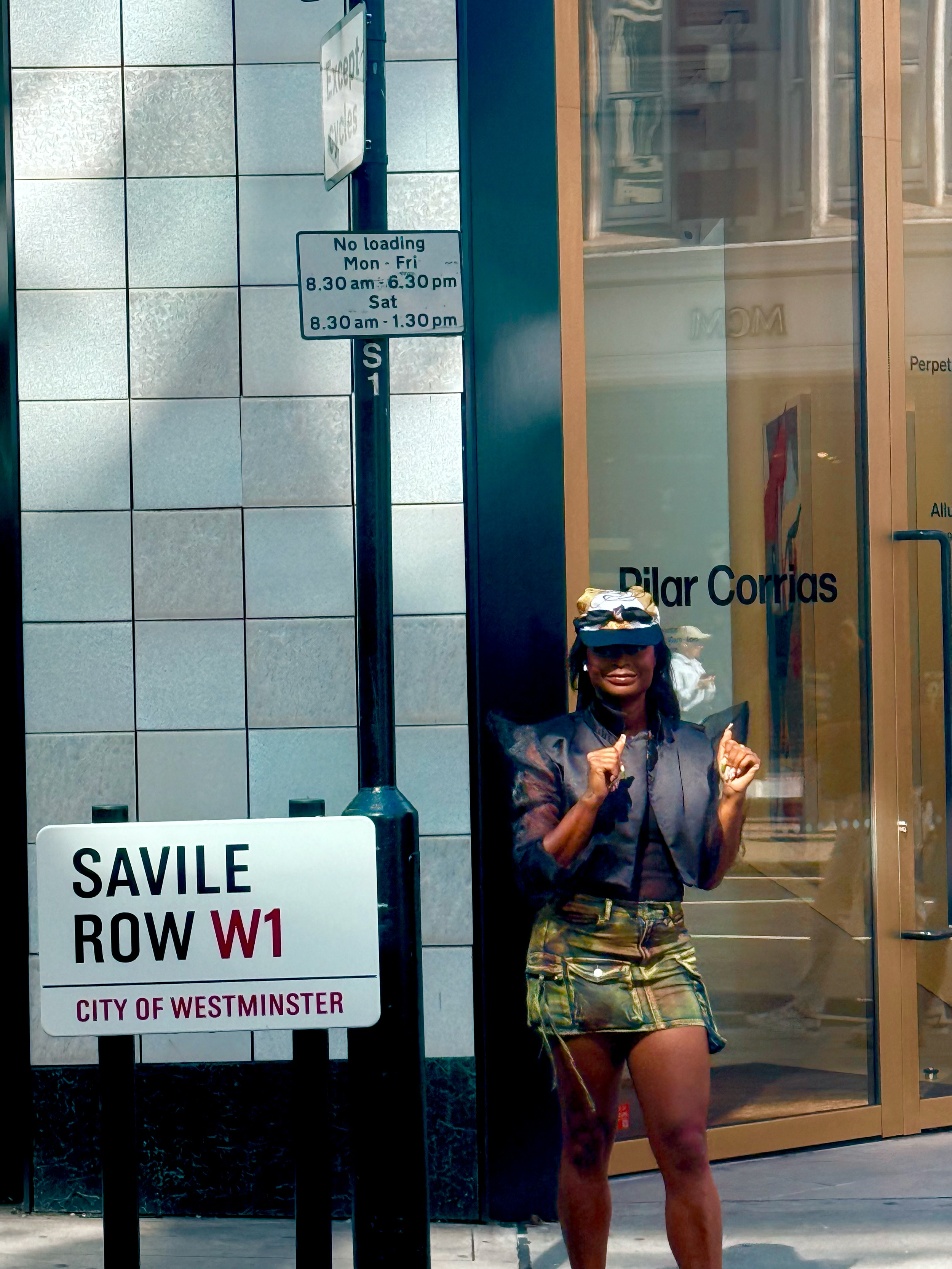 A woman standing outside a building with a sign for Savile Row W1 in Westminster, London, with a glass door to Pilar Corrías and a street sign indicating loading restrictions.