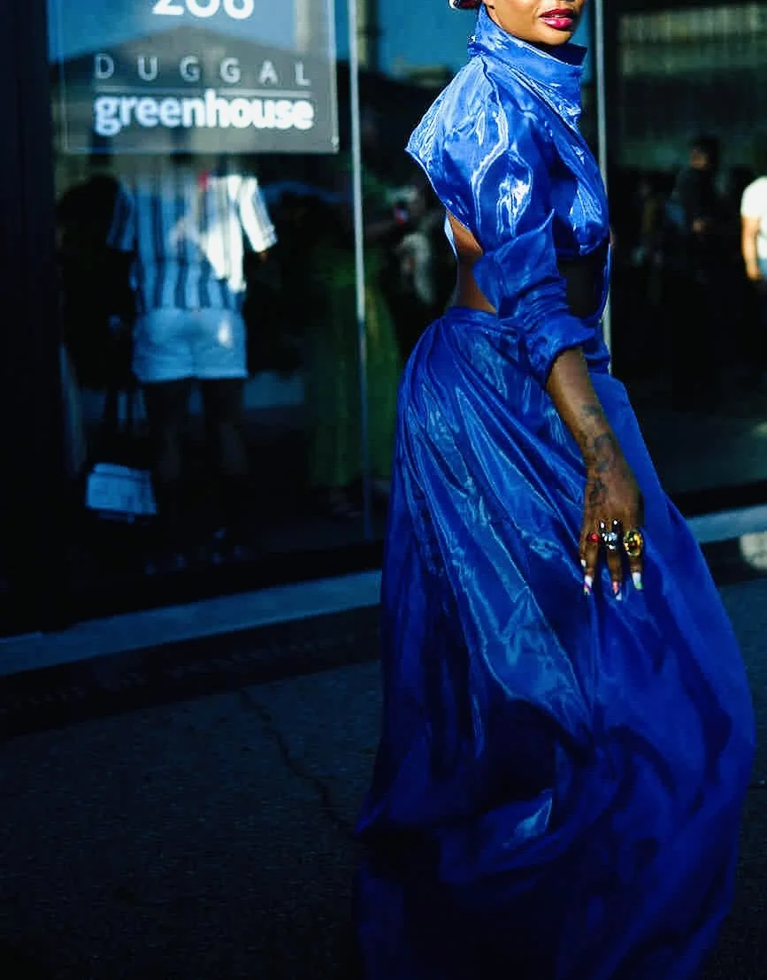 A woman in a shiny blue outfit standing outdoors near a glass building with store signs, including 'DUGGAL greenhouse.'