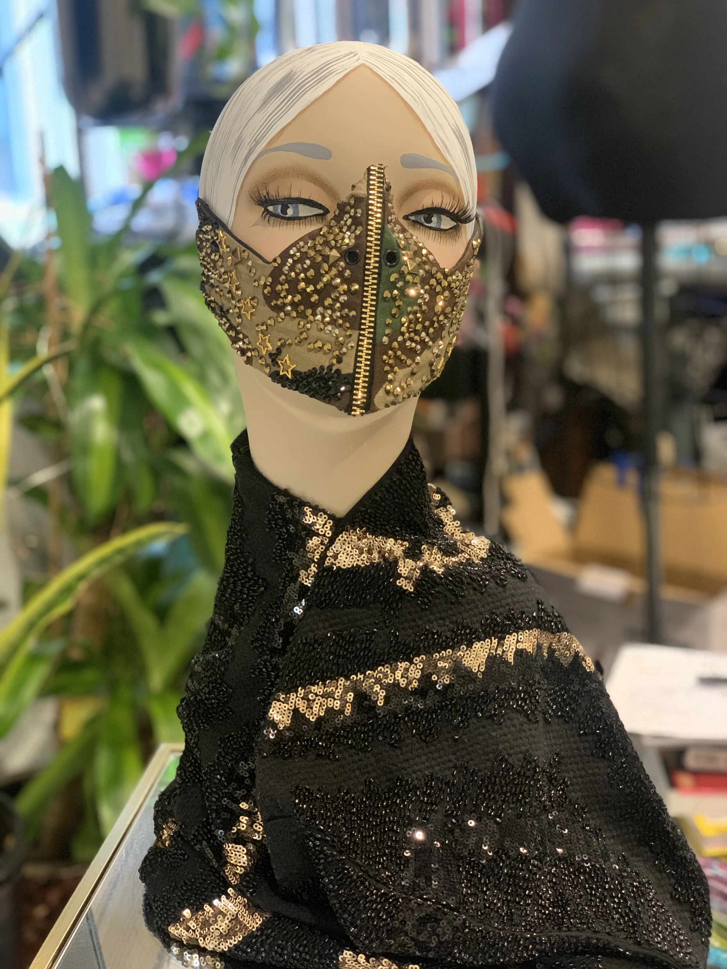 A mannequin head with platinum blonde hair and blue eyes, wearing a gold and black zippered face mask decorated with stars and rhinestones, and a black sequined sweater.