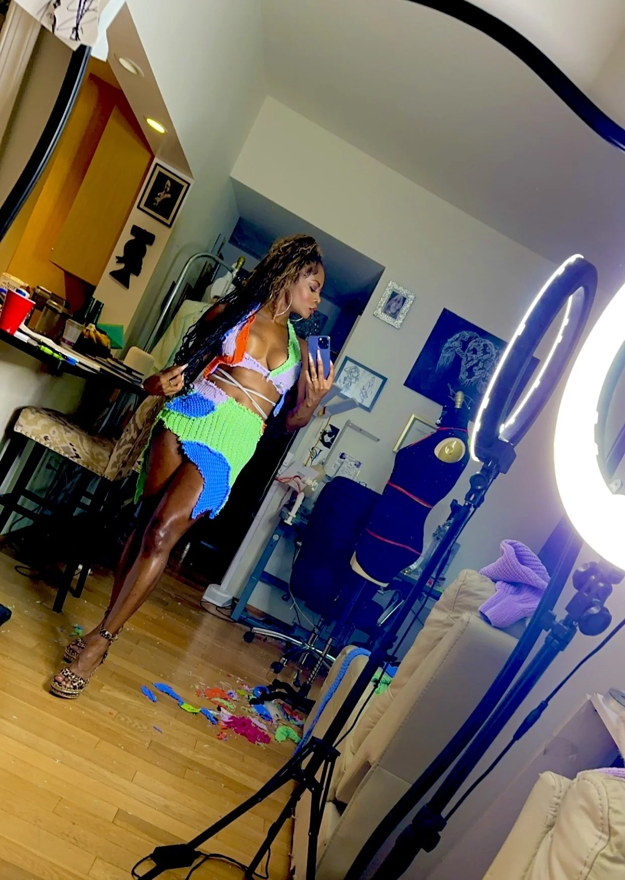 A woman taking a selfie in front of a mirror, wearing a colorful, revealing outfit with multicolored mesh pieces. The room has wooden floors, art on the walls, and photo equipment, with various objects scattered on the floor.