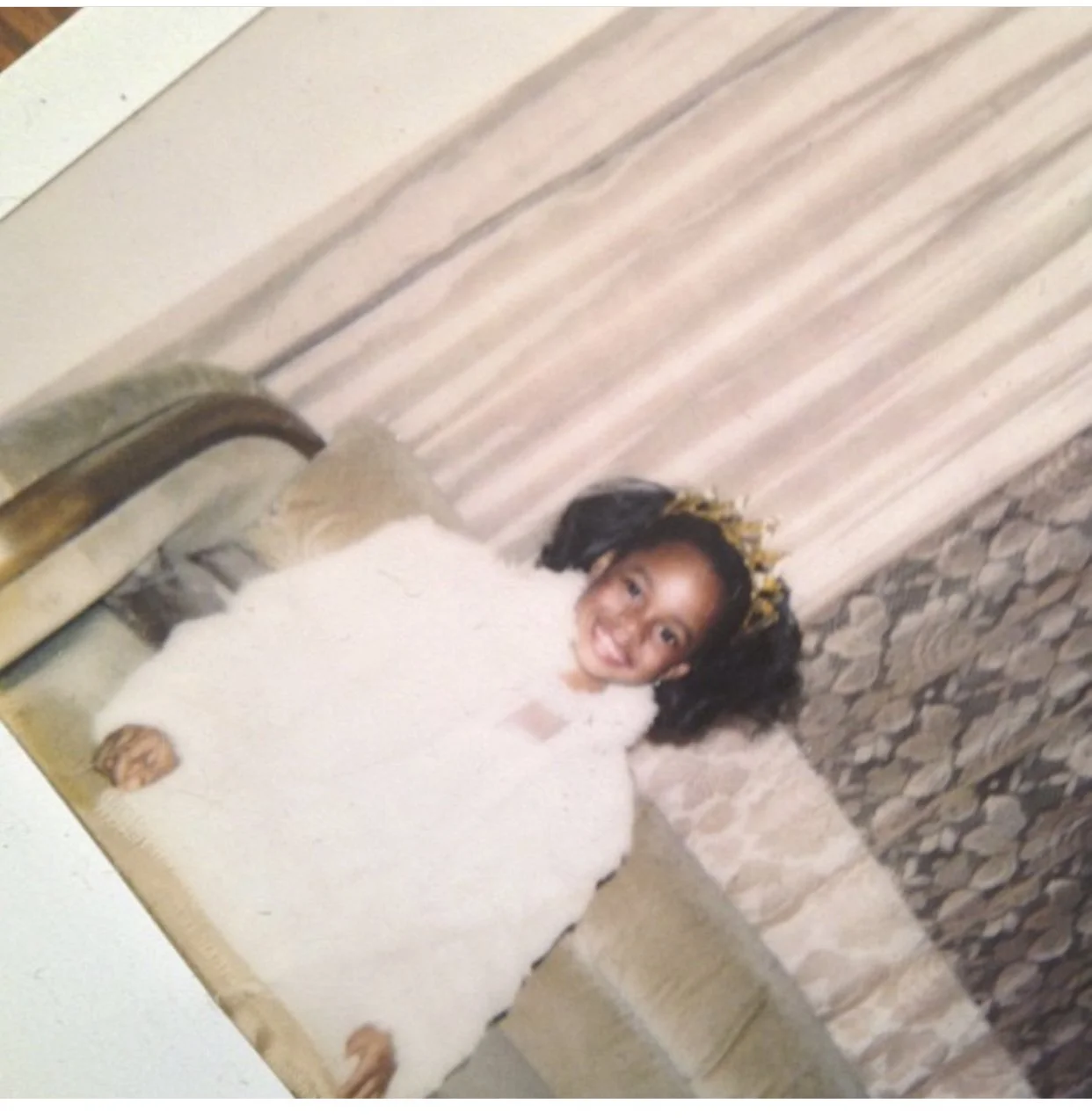 Young girl lying on a beige couch, smiling at the camera, wearing a white fluffy sweater and a yellow headband with black spots, in a living room with a brick wall and curtains in the background.